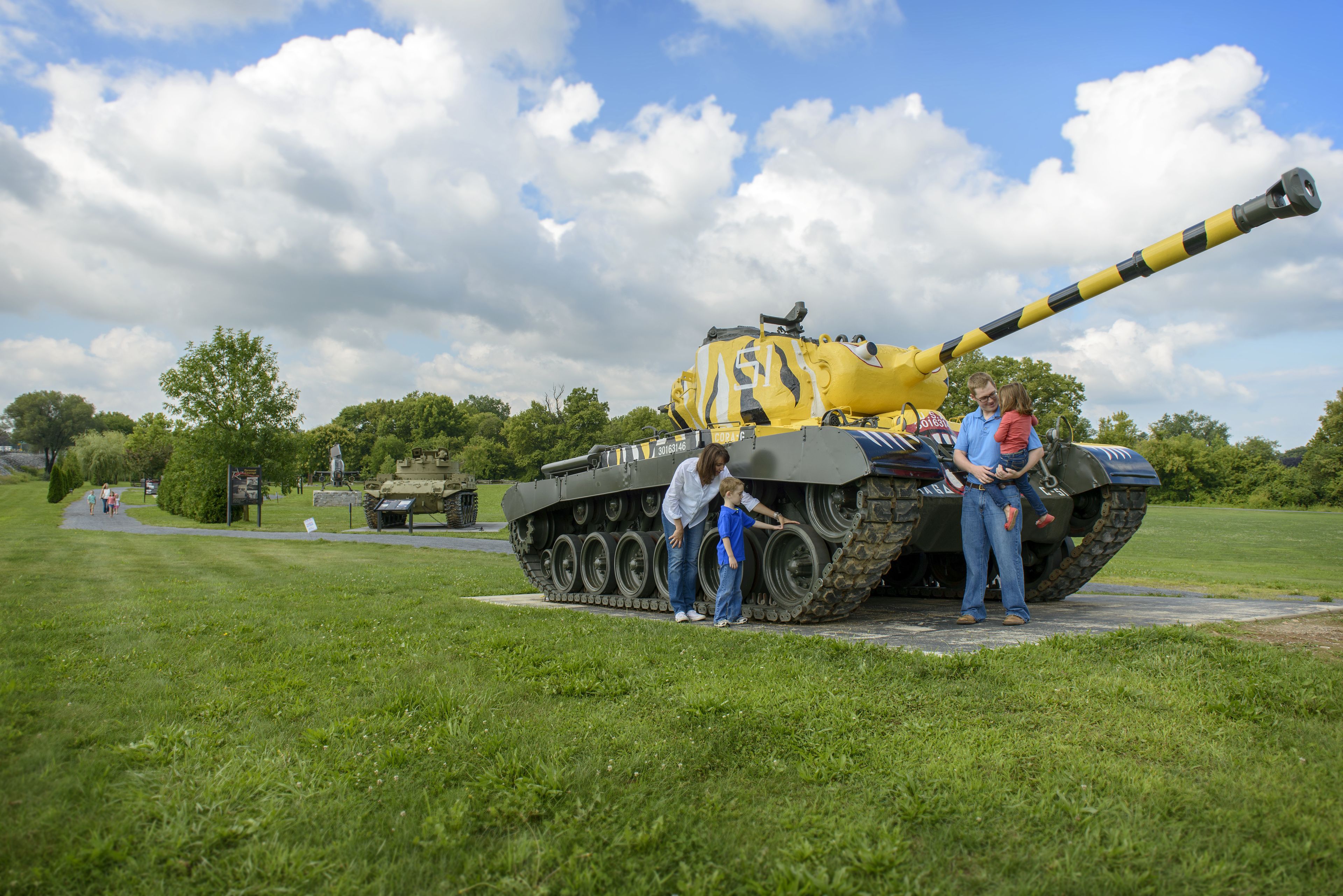 Un tanque militar exhibido en un campo abierto con una zona de césped y árboles al fondo.