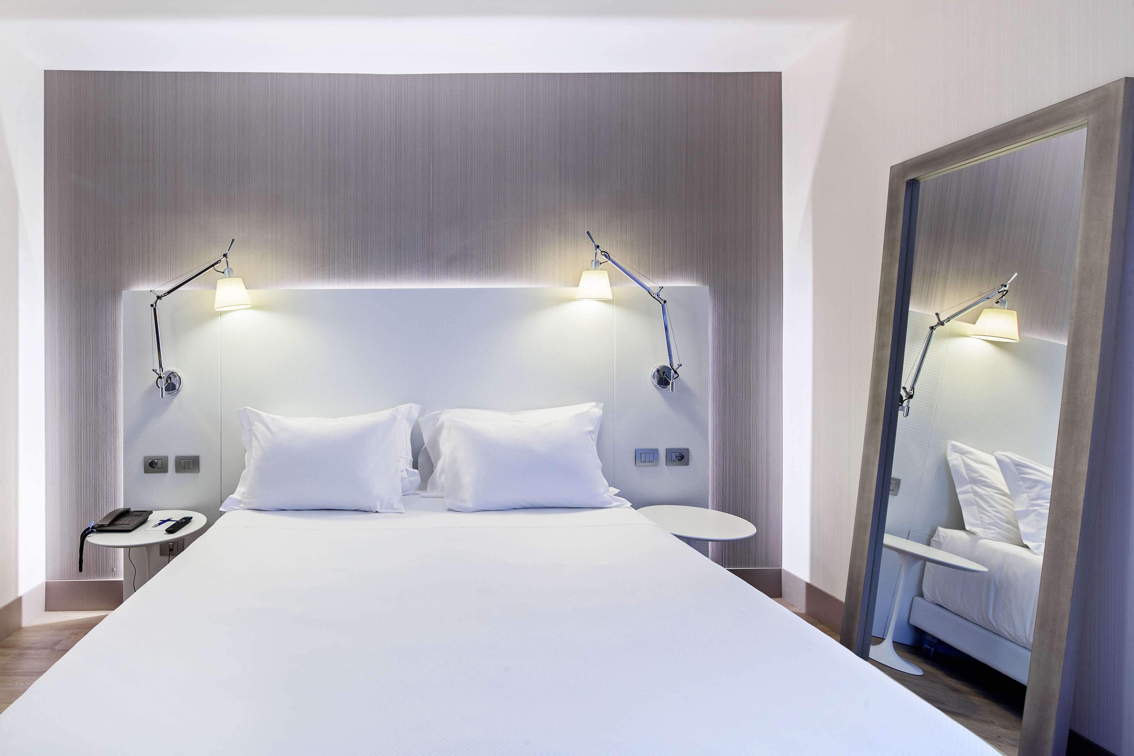 A modern hotel room with a large bed, two bedside tables, a small round table, and a mirror.