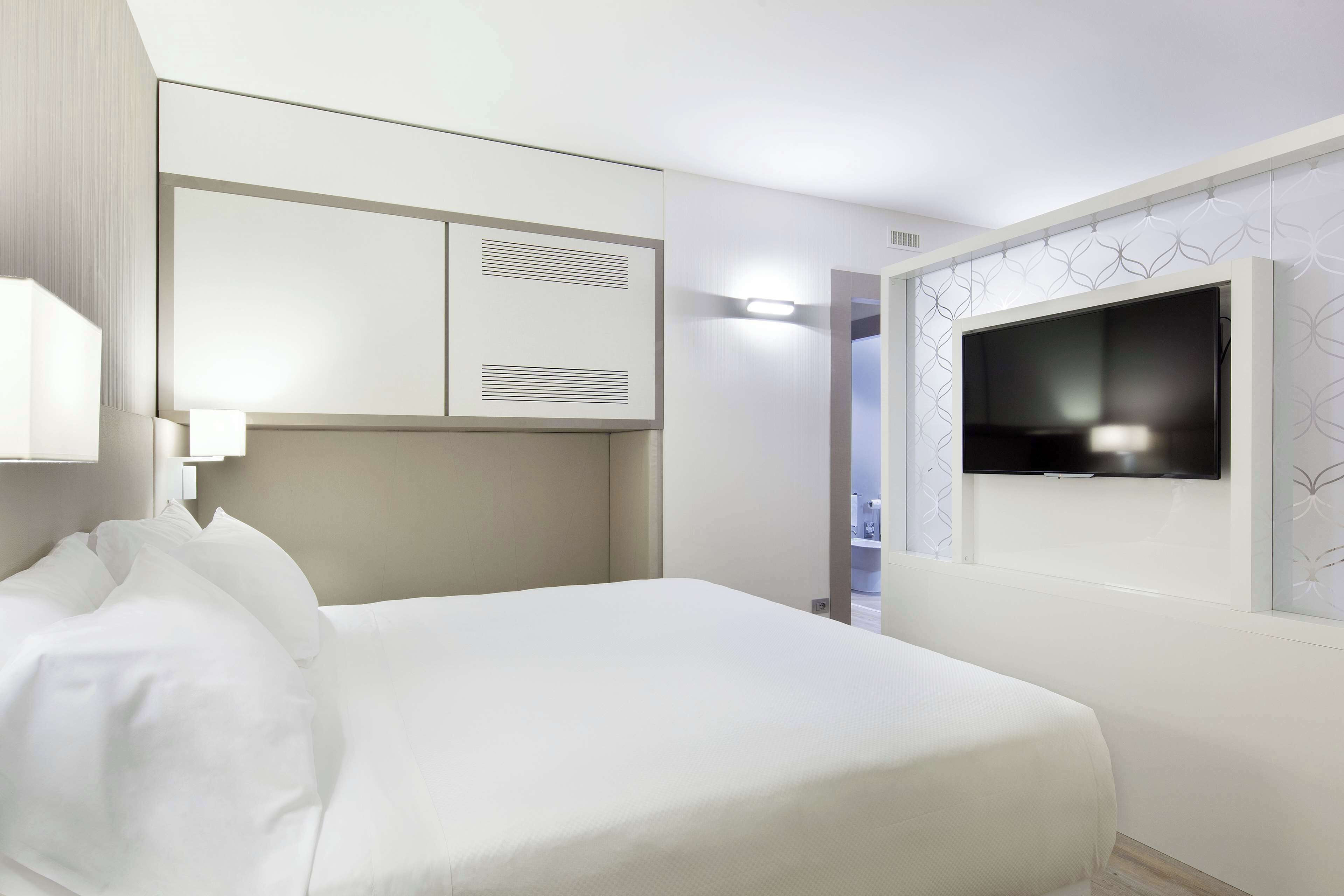A hotel room with a bed, a TV mounted on the wall, and a wardrobe with ventilation panels.