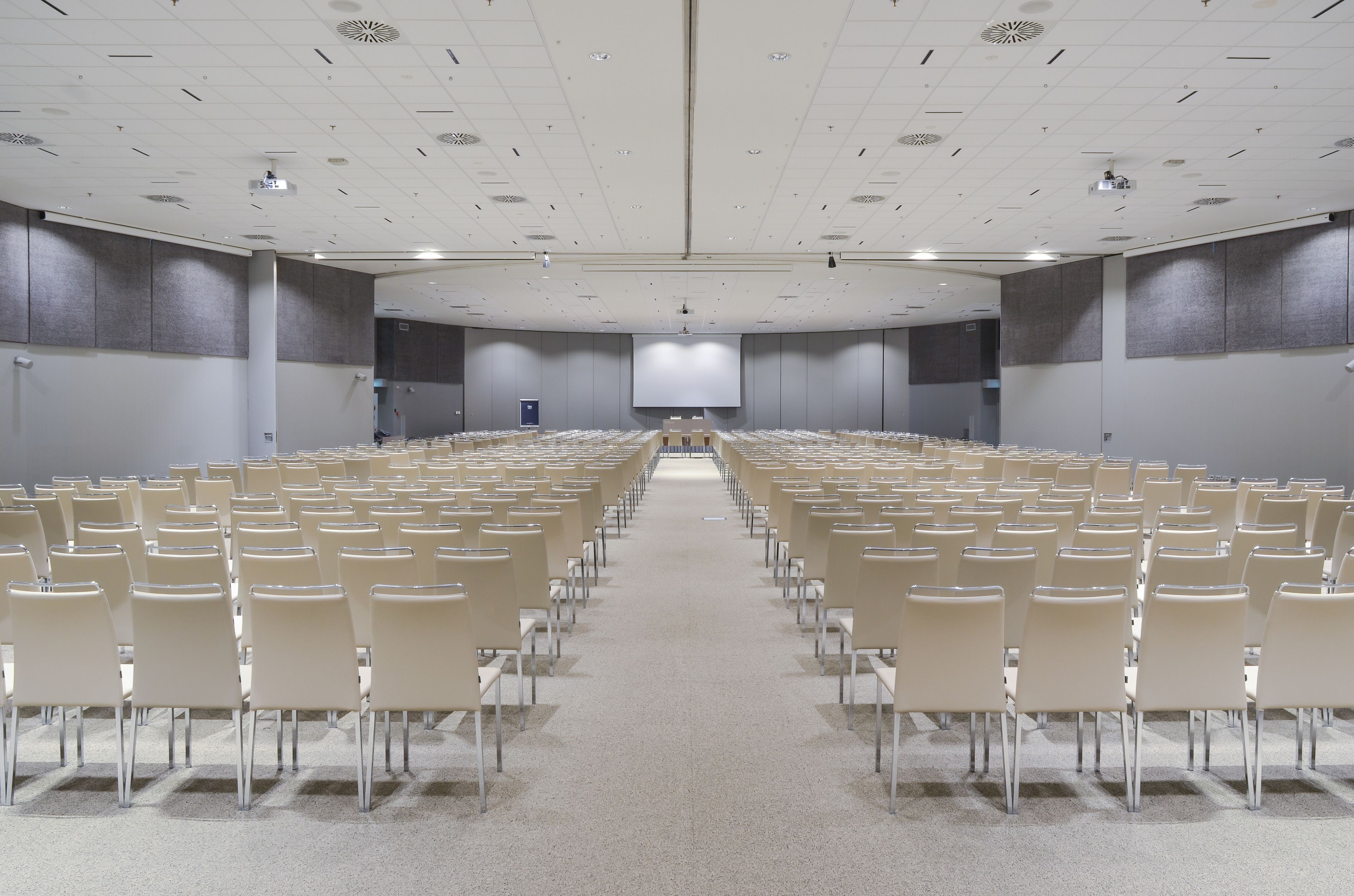 Un amplio salón de conferencias con filas de sillas orientadas hacia una pantalla central, equipado con proyectores de techo y altavoces.