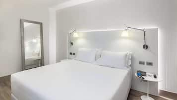 A modern hotel room with a large bed, two bedside lamps, a small table with a phone and remote, and a mirror on the wall.