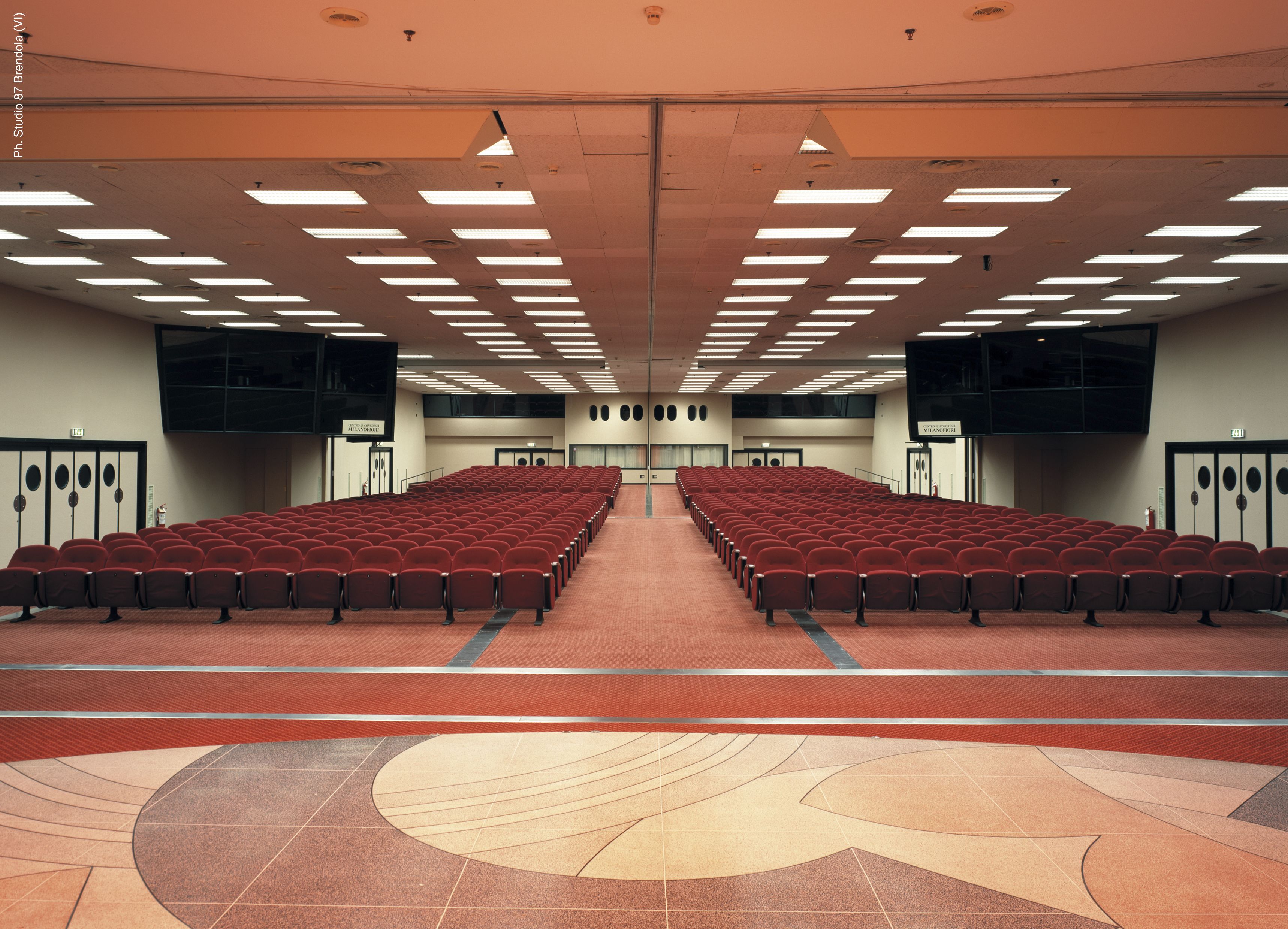 Un grand auditorium avec des rangées de sièges rouges, une scène à l’avant et un sol à motifs.