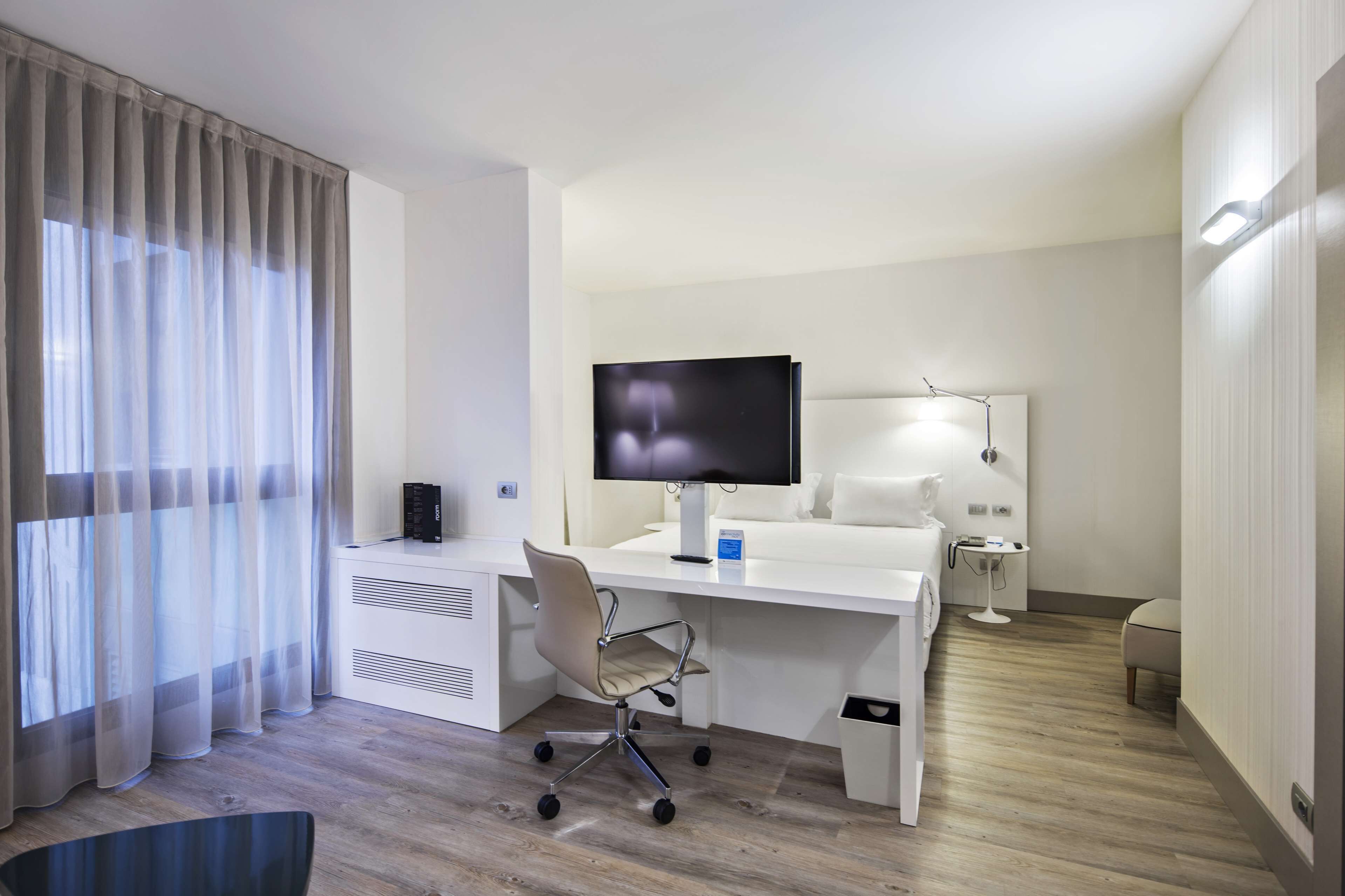 A modern hotel room with a white desk, a flat-screen TV, a chair, a bedside table with a lamp, and a waste bin.