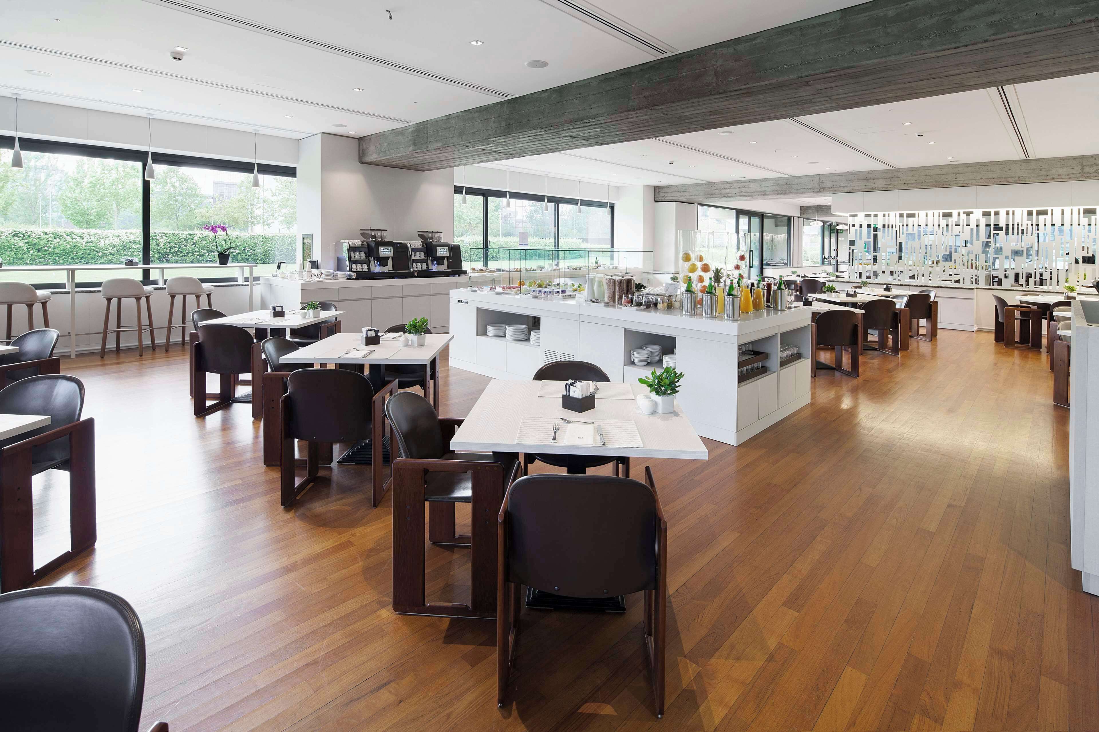 Ein modernes Restaurant mit Holzboden, weißen Tischen und schwarzen Stühlen. Es gibt eine Bar mit einer Theke und verschiedenen Flaschen.
