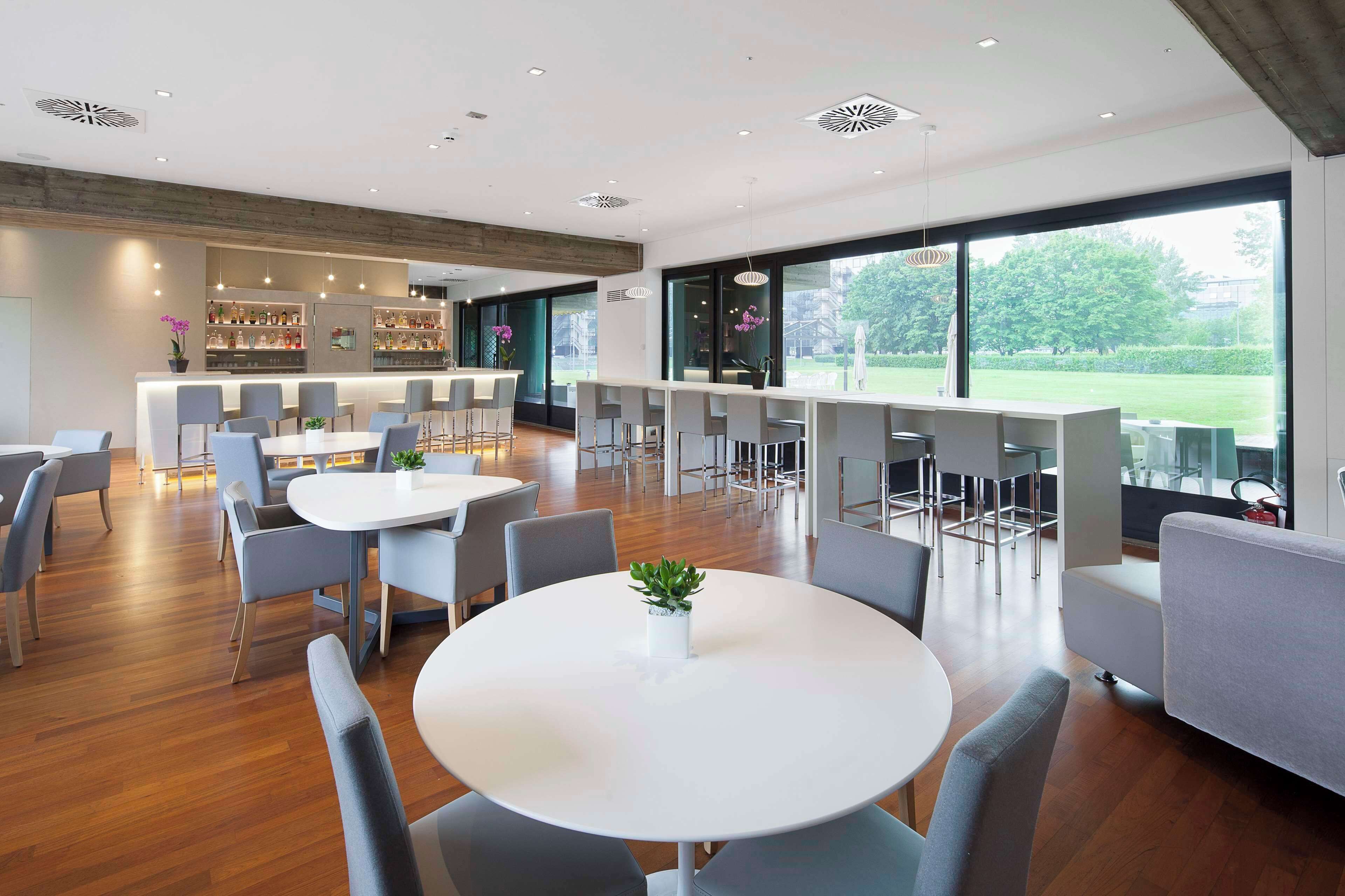 Un espace repas moderne avec des tables rondes et des chaises grises, un bar avec des étagères remplies de bouteilles et de grandes fenêtres offrant une vue sur l’extérieur.