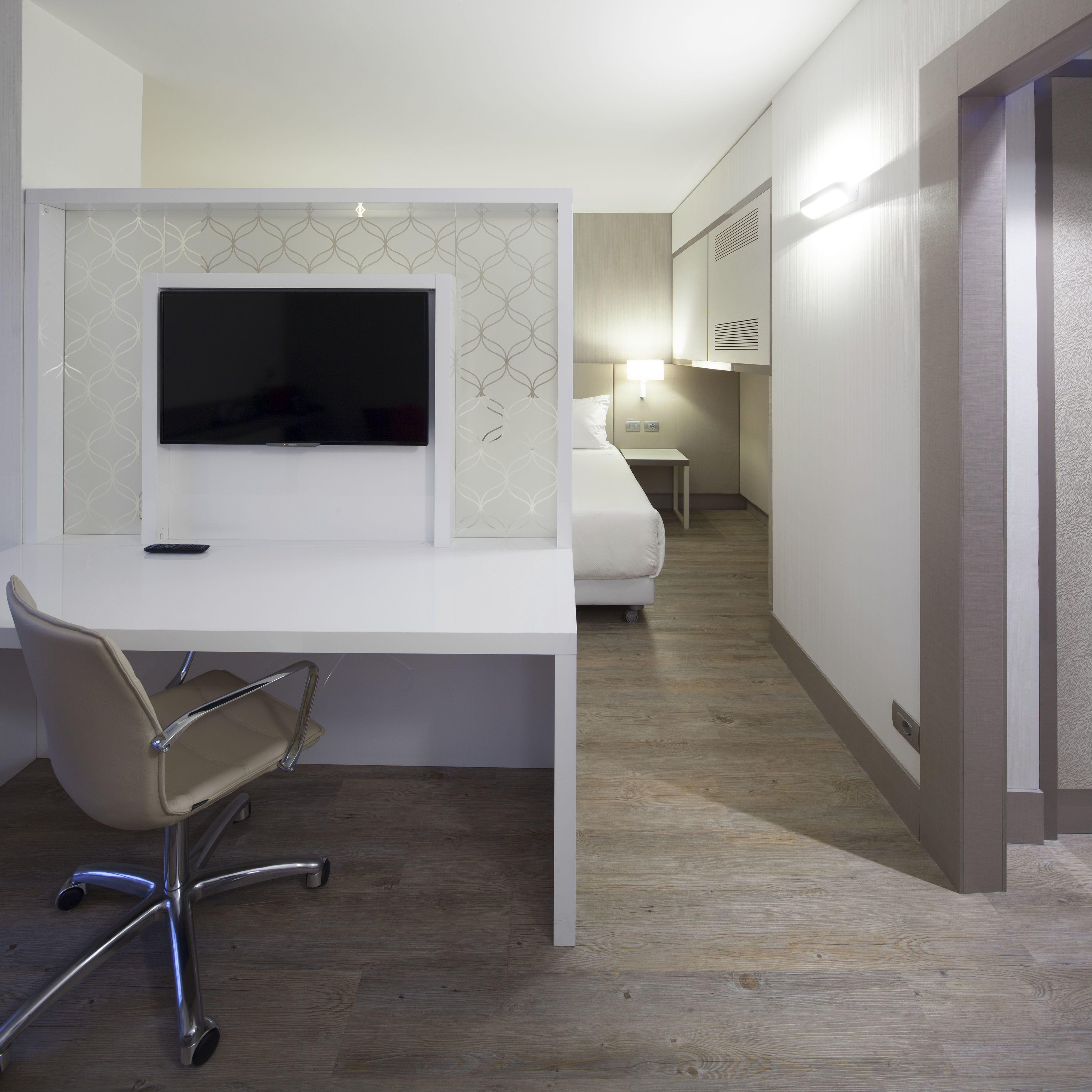 A modern hotel room with a white desk, a TV, a chair, a bedside table, and a bed.