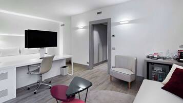 A modern hotel room with a flat-screen TV, a desk, a chair, a sofa, and a minibar.