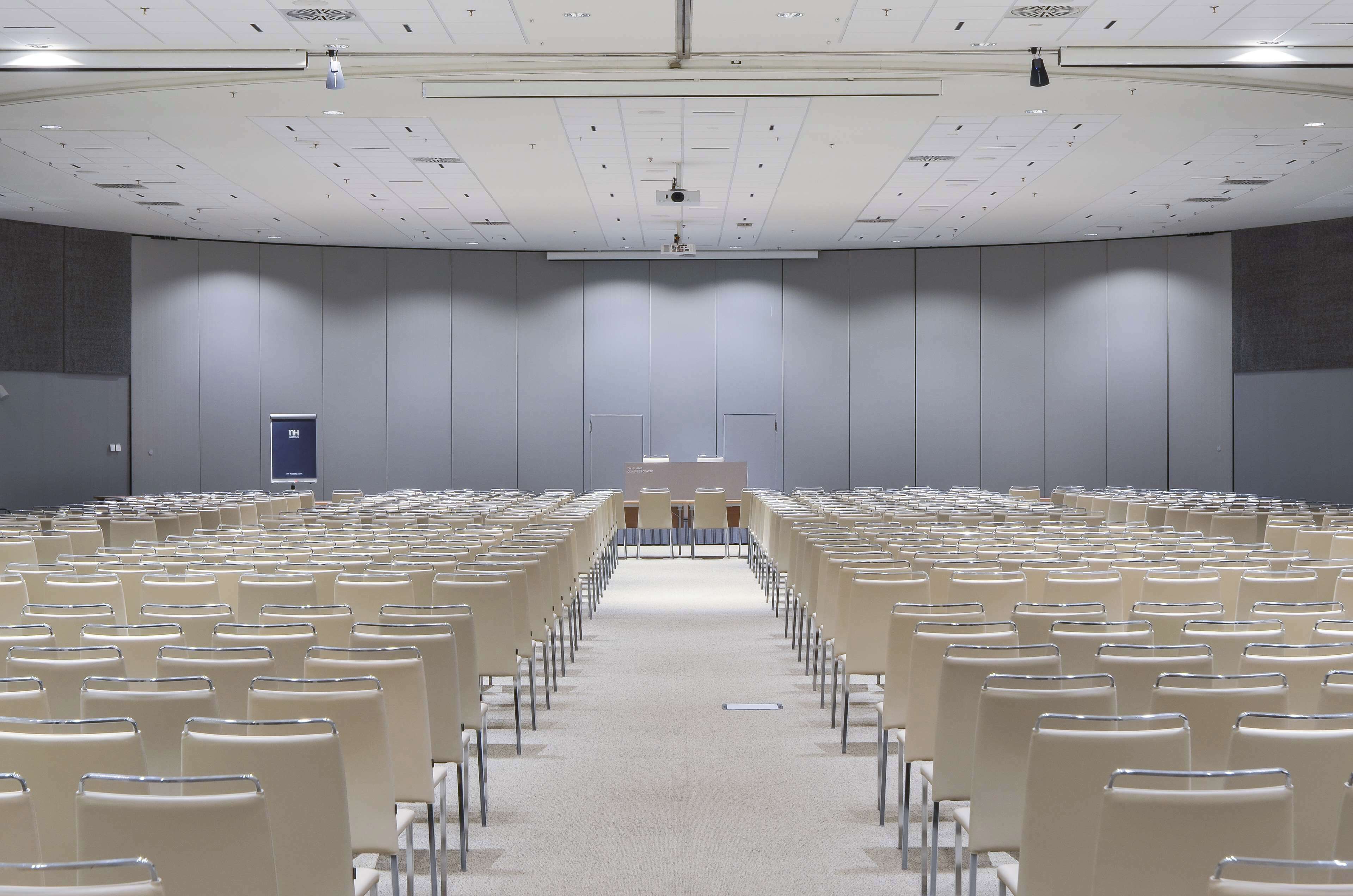 Un amplio salón de conferencias con filas de sillas, un atril y una pantalla de proyección.