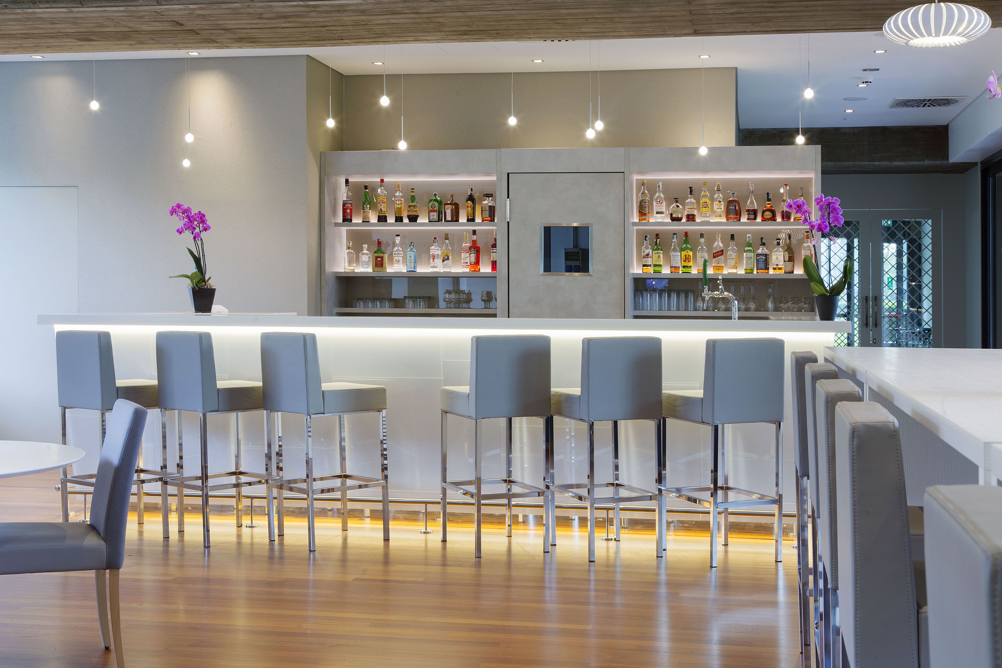 Uma área de bar moderna com bancada branca, banquetas e prateleiras cheias de garrafas.
