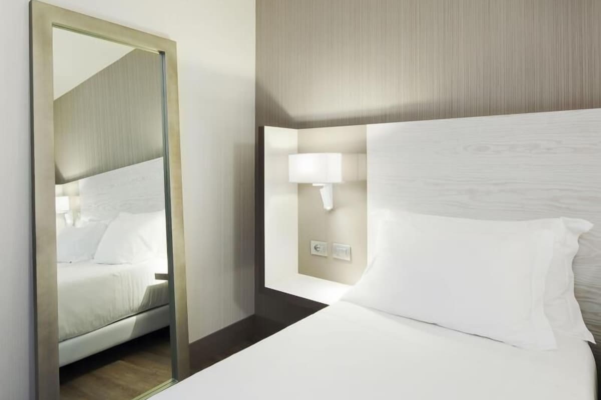 Una habitación de hotel con cama, mesita de noche, lámpara de pared y espejo.