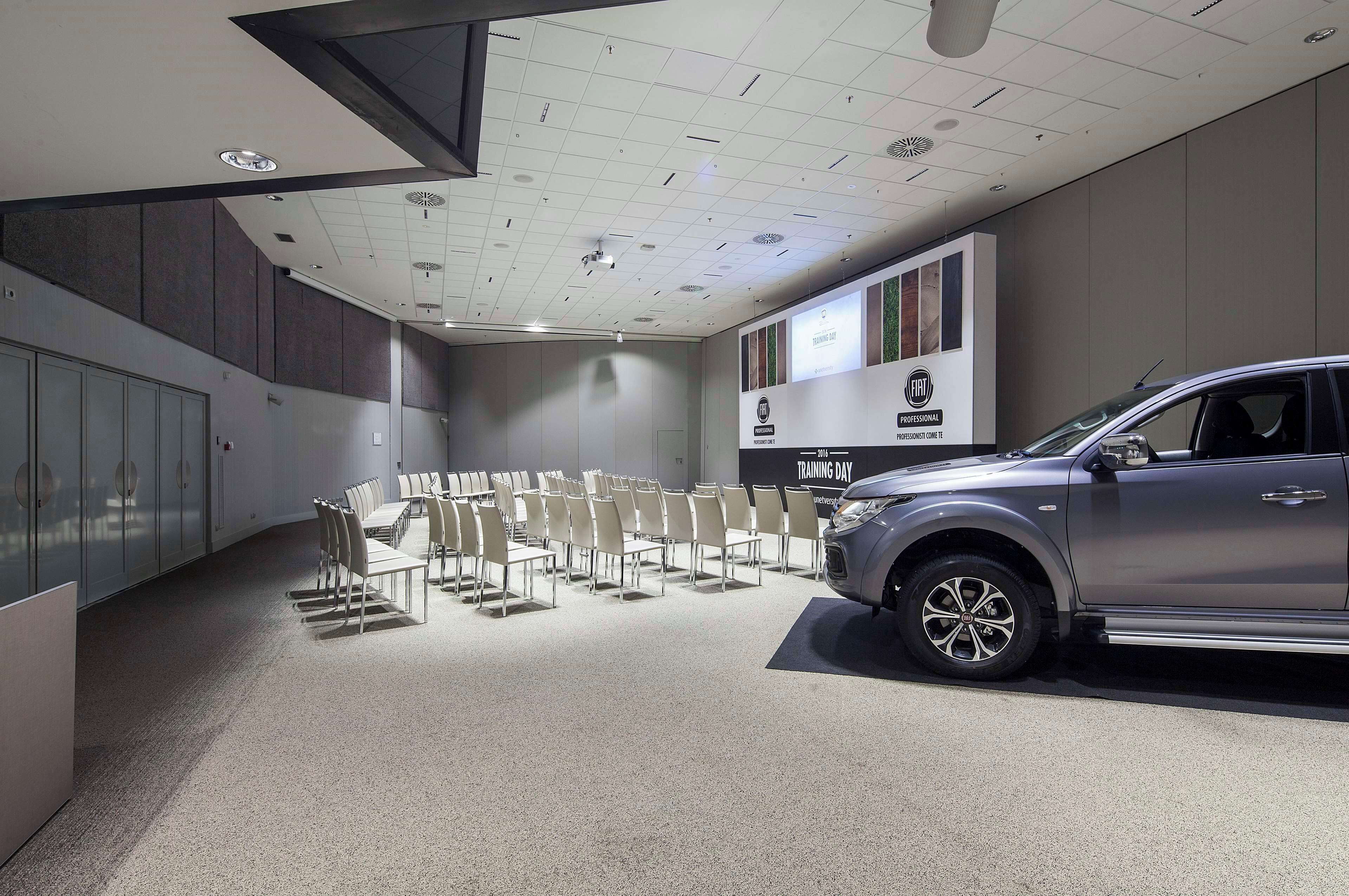 A conference room with a grey SUV on display, white chairs arranged in rows, and a white ceiling with recessed lighting.