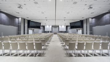 Un salón de conferencias con filas de sillas, una pantalla grande y un proyector.