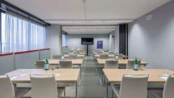 Sala de conferências com mesas, cadeiras, uma televisão e garrafas de água.