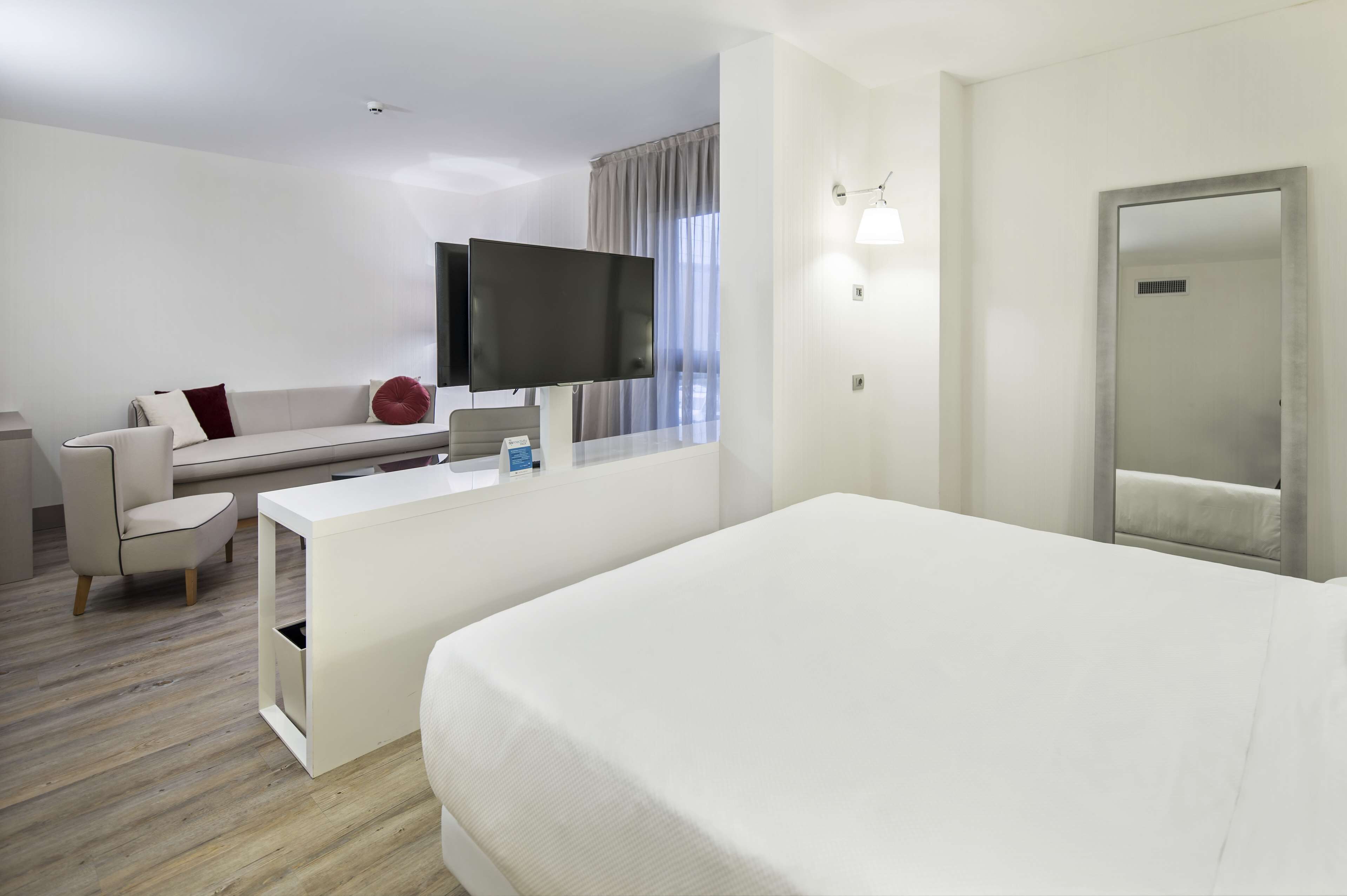 A modern hotel room with a large bed, a desk with a flat-screen TV, a sofa, and a large mirror.