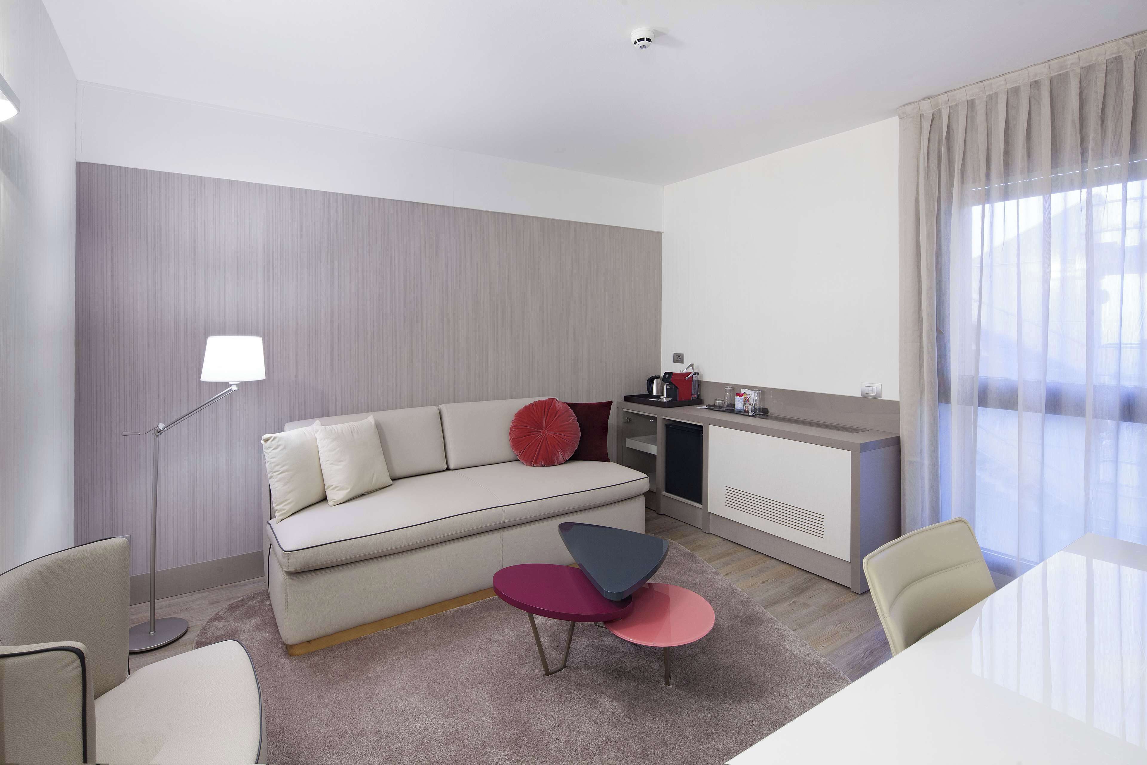 A modern hotel room with a sofa, a small table, a lamp, a kitchenette, and a large window with curtains.