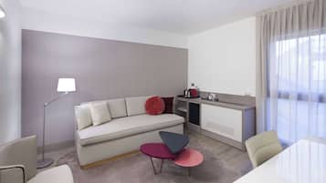 A modern hotel room with a sofa, a small table, a lamp, a kitchenette, and a large window with curtains.