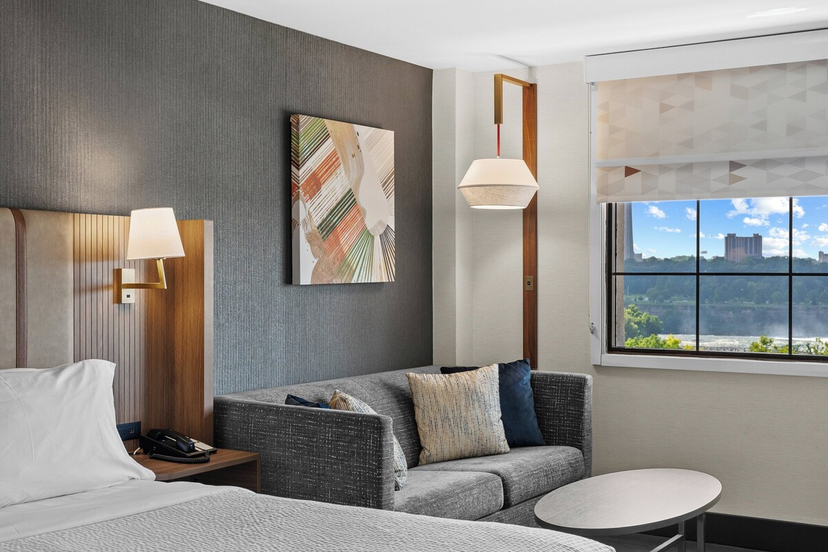 A modern hotel room with a grey sofa, a small round table, a bed with white bedding, a painting on the wall, and a window with a city view.
