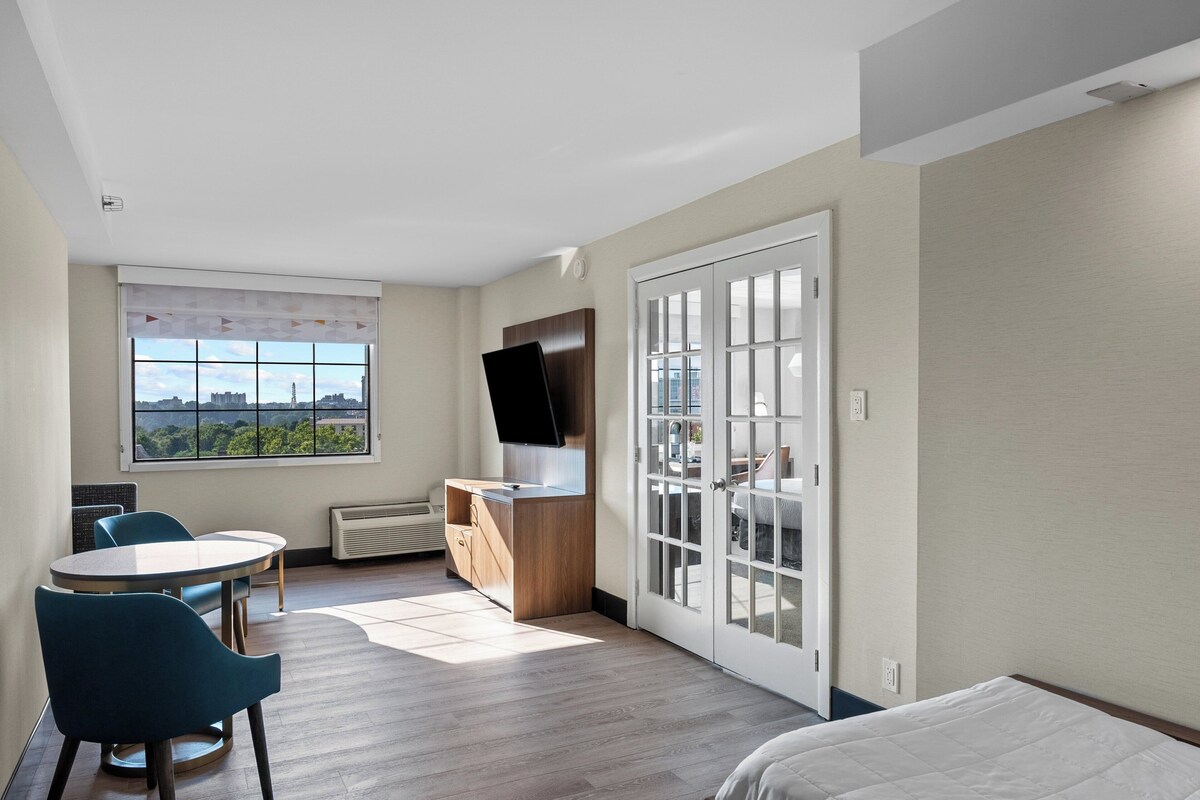 A hotel room with a bed, a desk with a flat-screen TV, a dining table with chairs, and a large window with a city view.