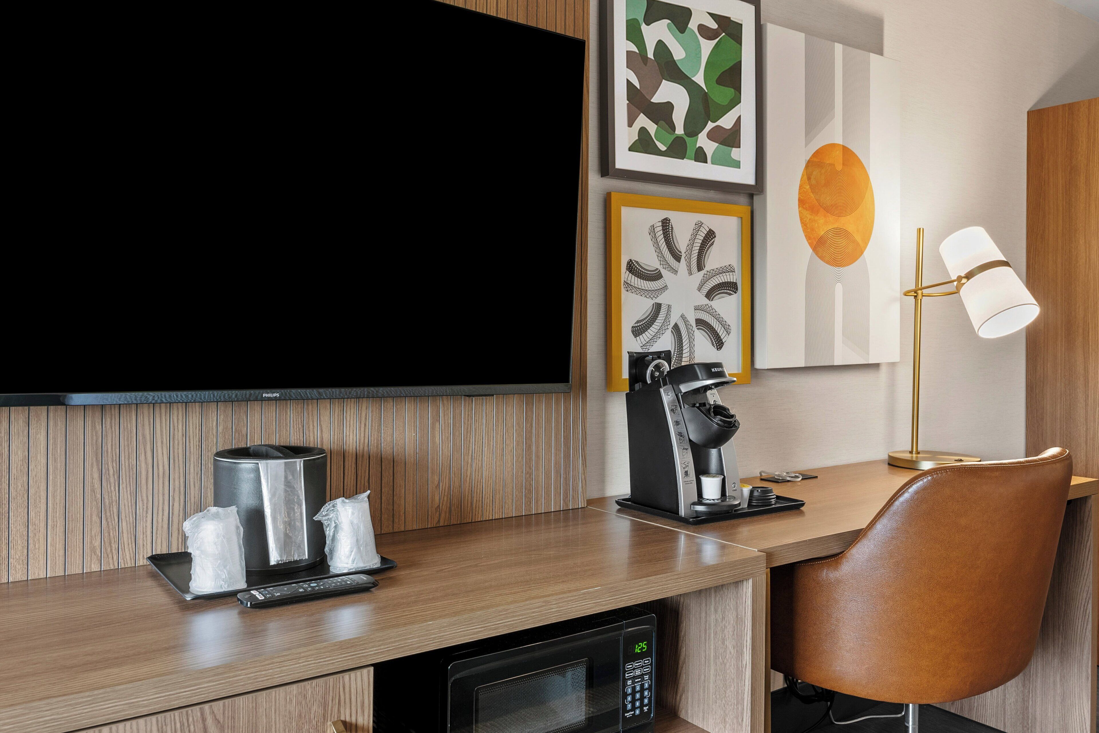 A modern hotel room with a wooden desk, a television, a coffee machine, a microwave, and a leather chair.