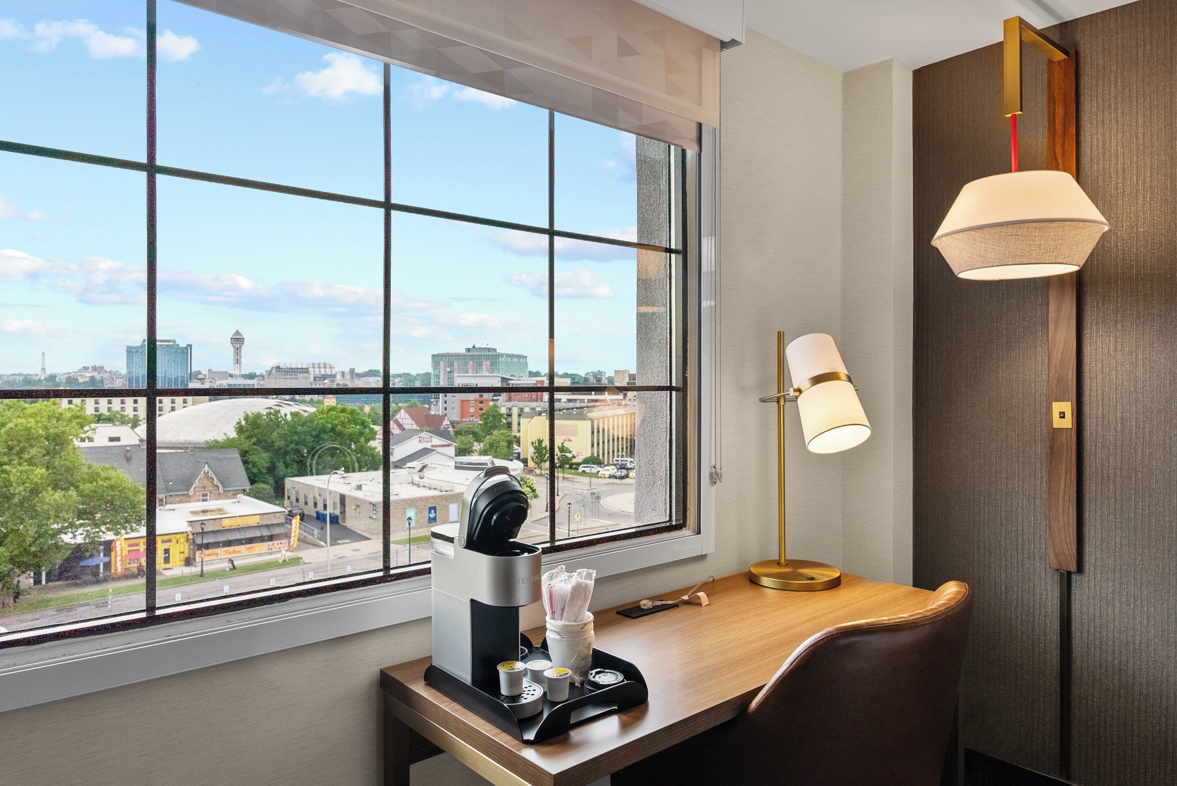 Une chambre d’hôtel dotée d’une grande fenêtre donnant sur la ville, d’un bureau avec une machine à café, une lampe et une chaise.