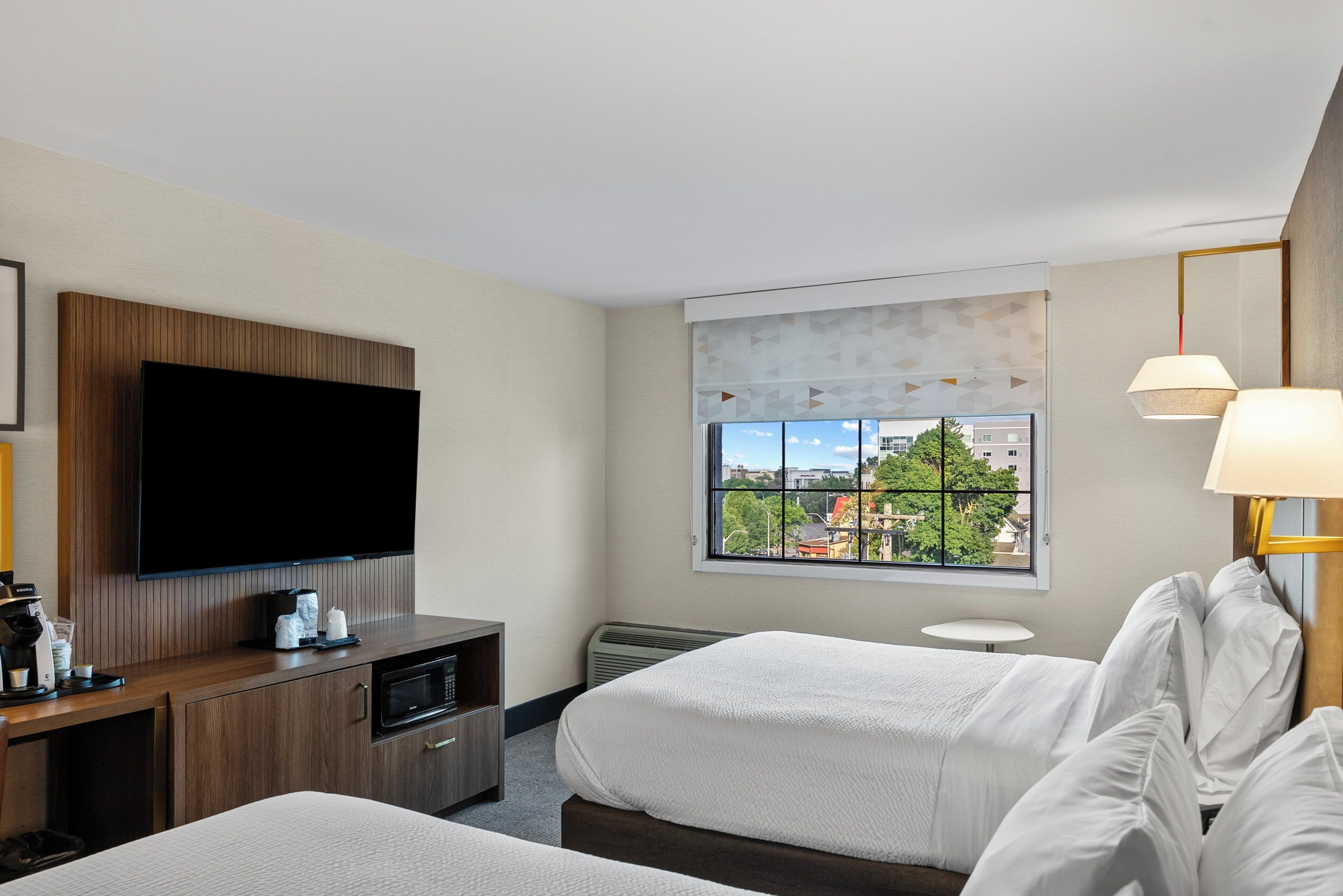 A hotel room with a large flat-screen TV, a wooden dresser, a bed with white bedding, a small round table, and a window with a city view.