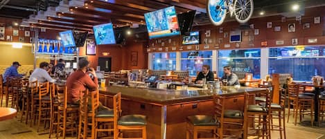 A bar area with wooden stools, a bar counter, and multiple screens displaying content.