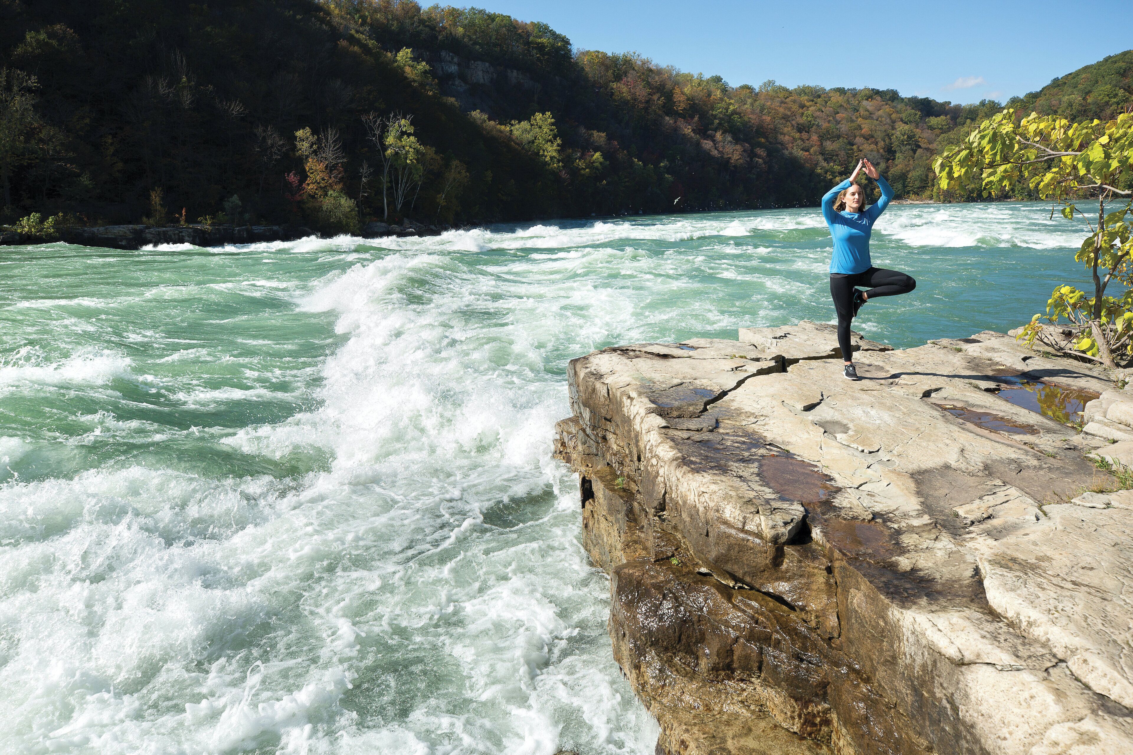 A person performing yoga on a rocky ledge by a river.