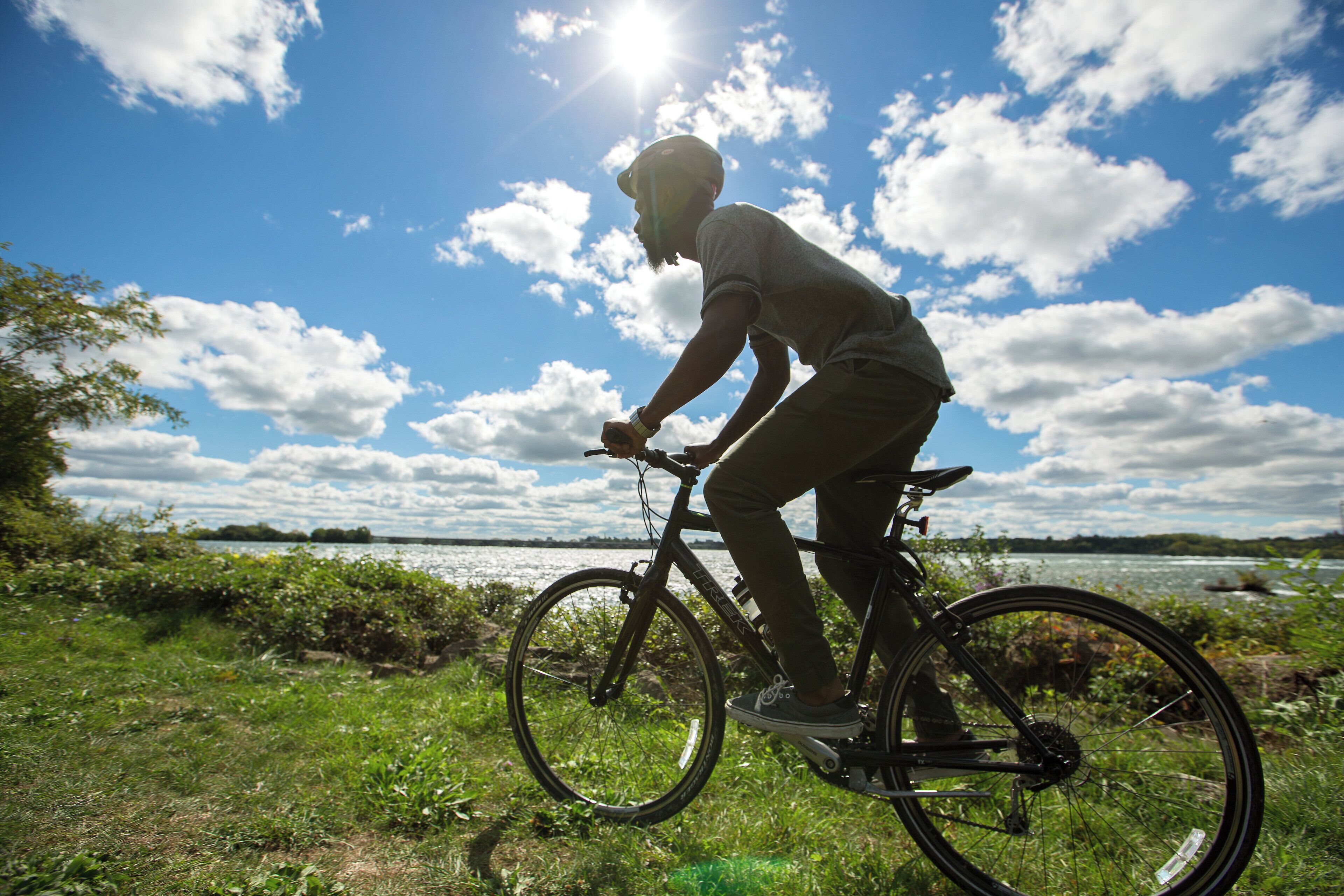 A person riding a bicycle on a grassy path with a body of water and a cloudy sky in the background.