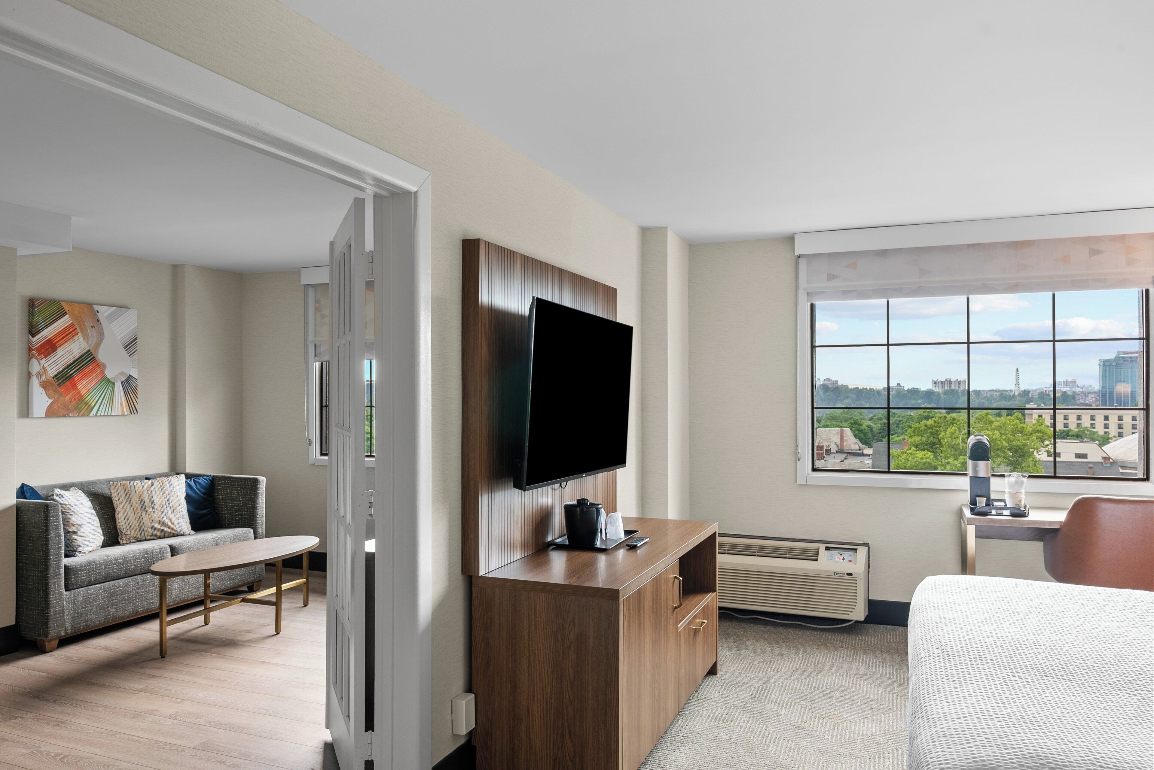 Une chambre d’hôtel comprenant un canapé, une table basse, un téléviseur fixé sur un meuble en bois, une grande fenêtre donnant sur la ville et un lit.