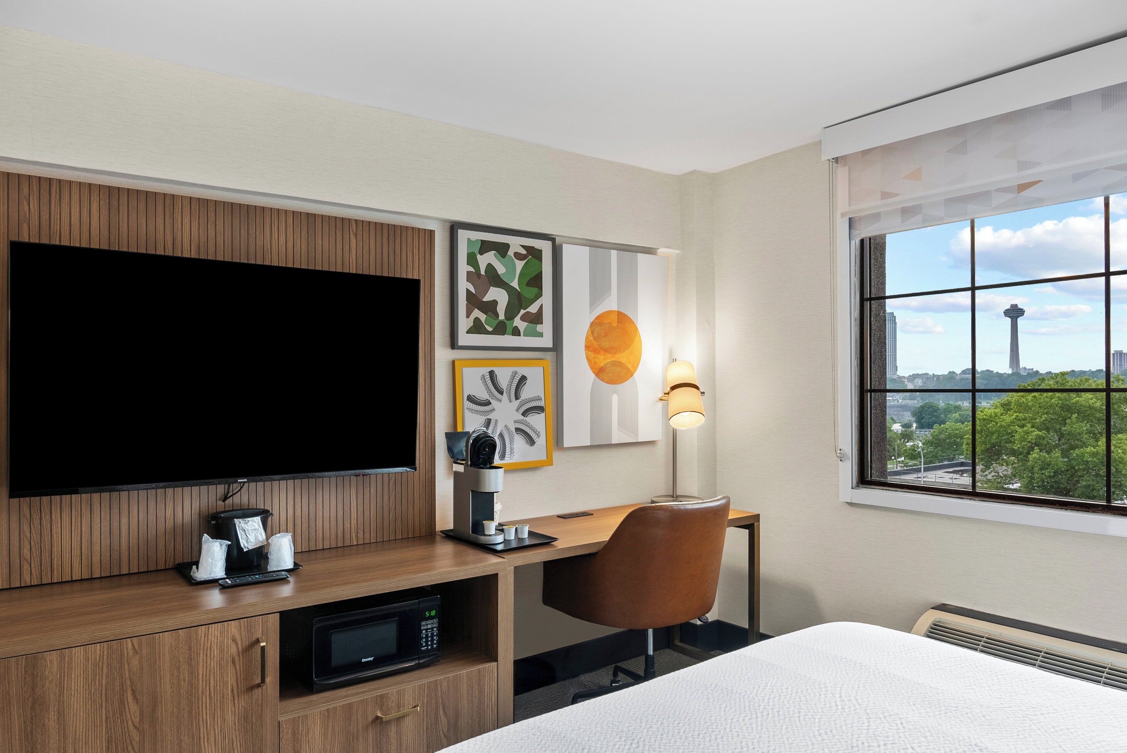 A hotel room with a large flat-screen TV, a desk with a coffee maker, a chair, and a bed.