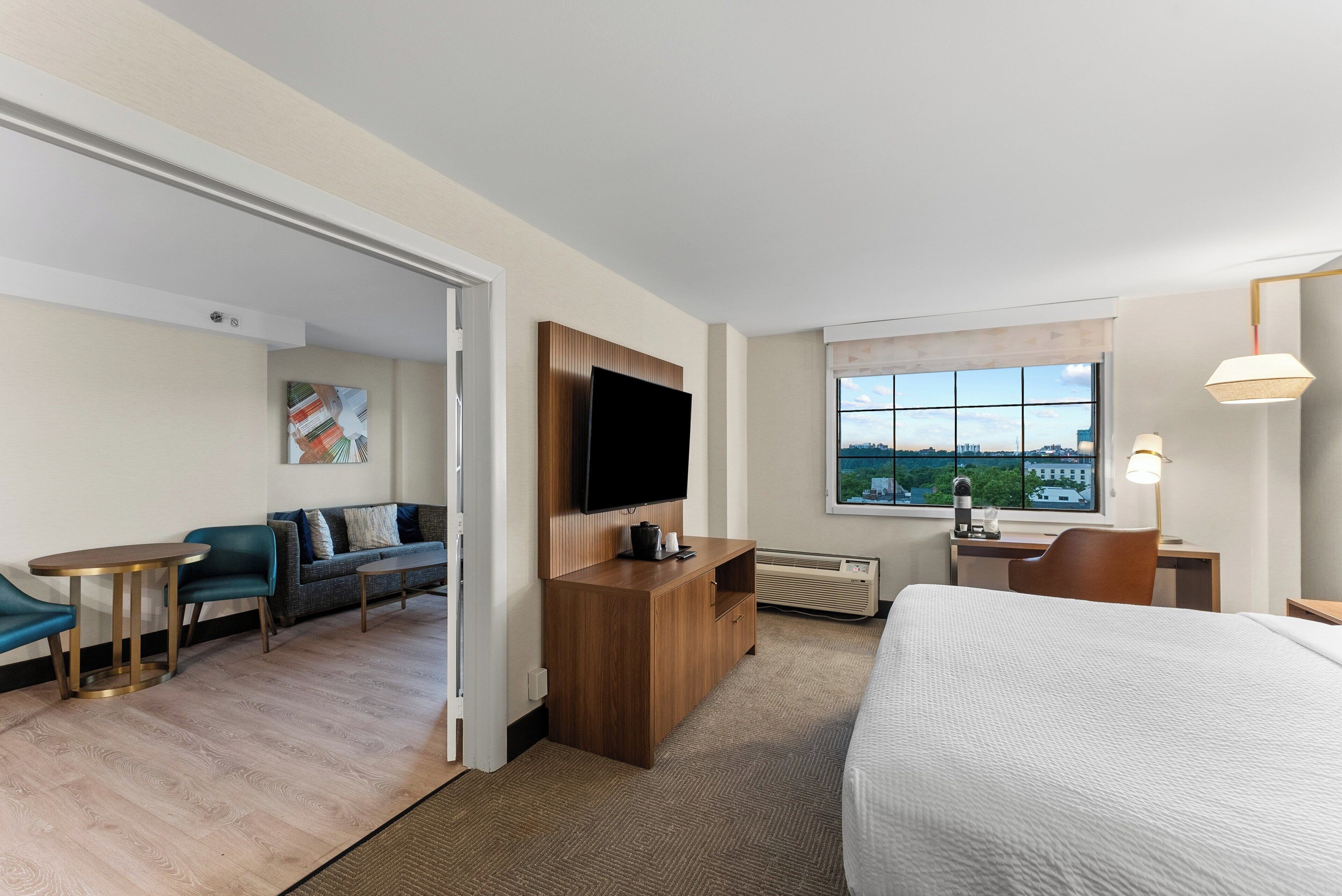 A hotel room with a bed, a TV, a desk, a chair, a sofa, and a window with a city view.