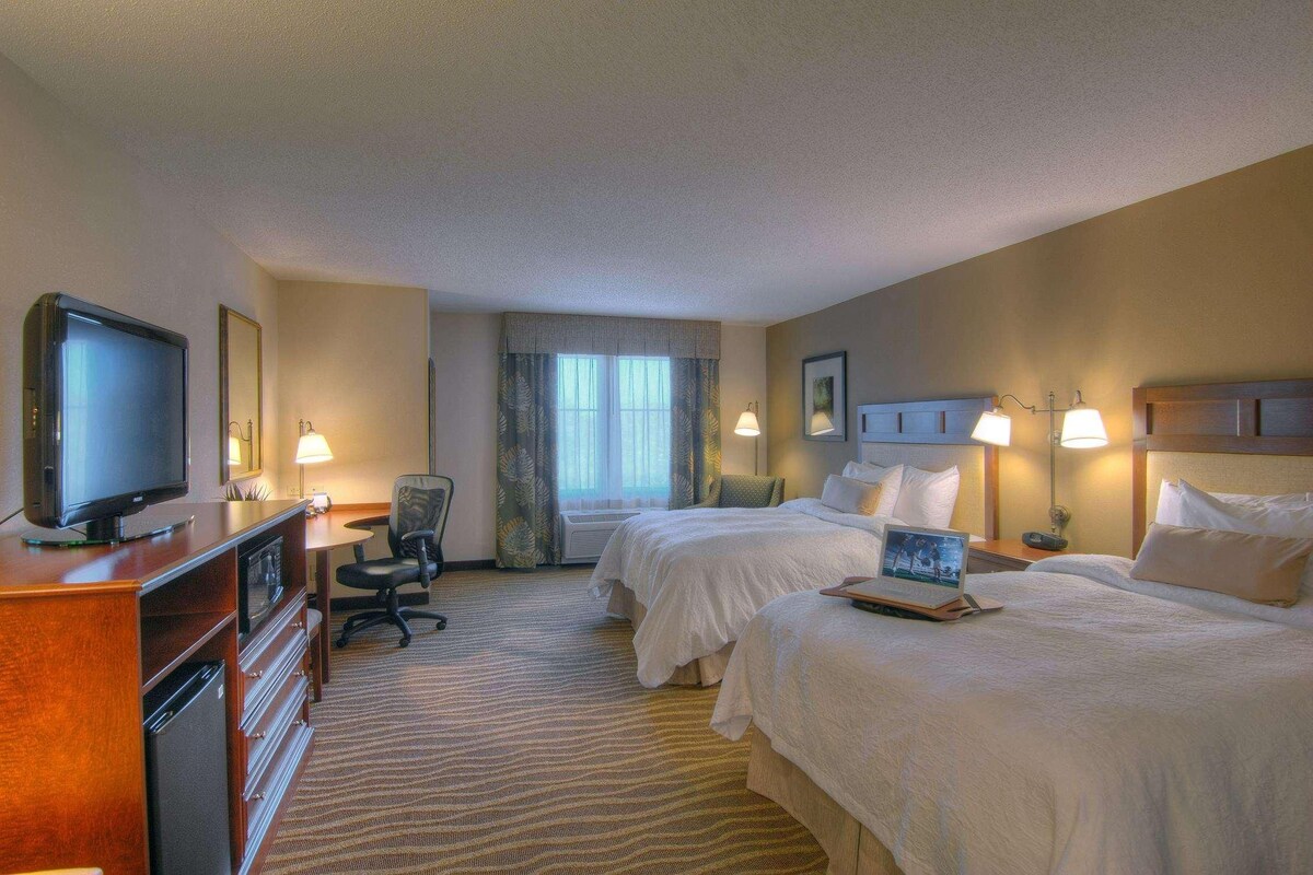 A hotel room with two beds, a desk with a television, a laptop, and a chair.