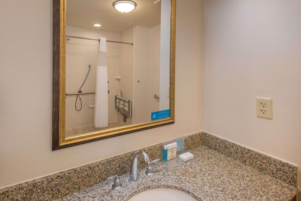 A bathroom with a granite countertop, a sink, a mirror with a gold frame, a towel rack, and a shower area.