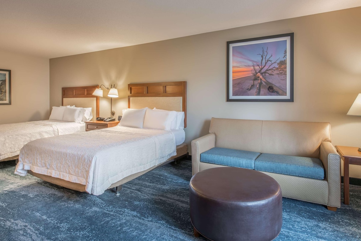 Habitación de hotel con dos camas, un sofá, un taburete redondo, una mesita de noche con lámparas y un cuadro en la pared.