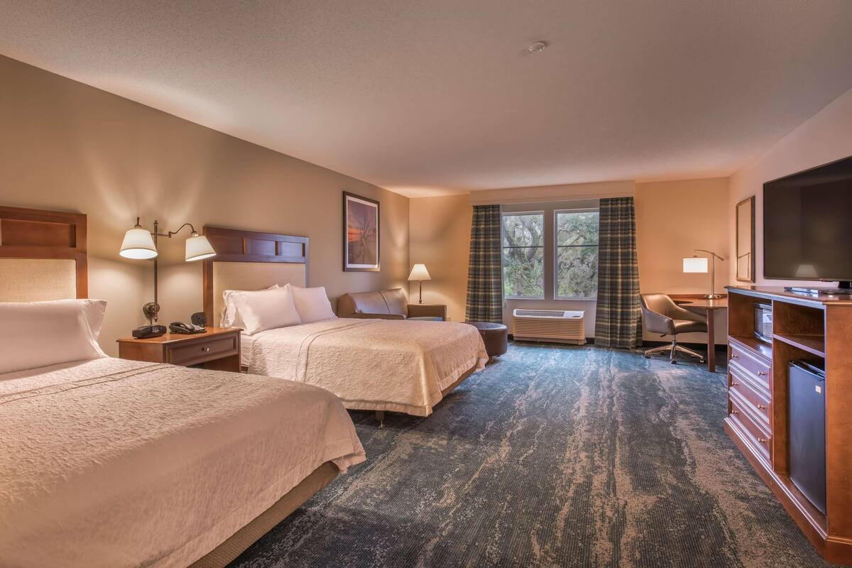 A hotel room with two beds, a desk, a television, and a window with curtains.