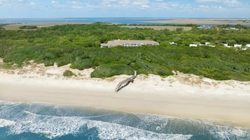 An aerial view of a coastal area with a beach, a wooden boardwalk, and a large building surrounded by dense greenery.