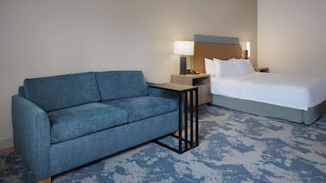 A hotel room with a blue sofa, a bed with white bedding, a nightstand with a lamp, and a patterned carpet.