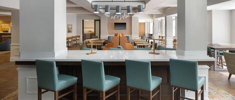 A modern hotel lobby with a central bar area, seating arrangements, and a large television screen.