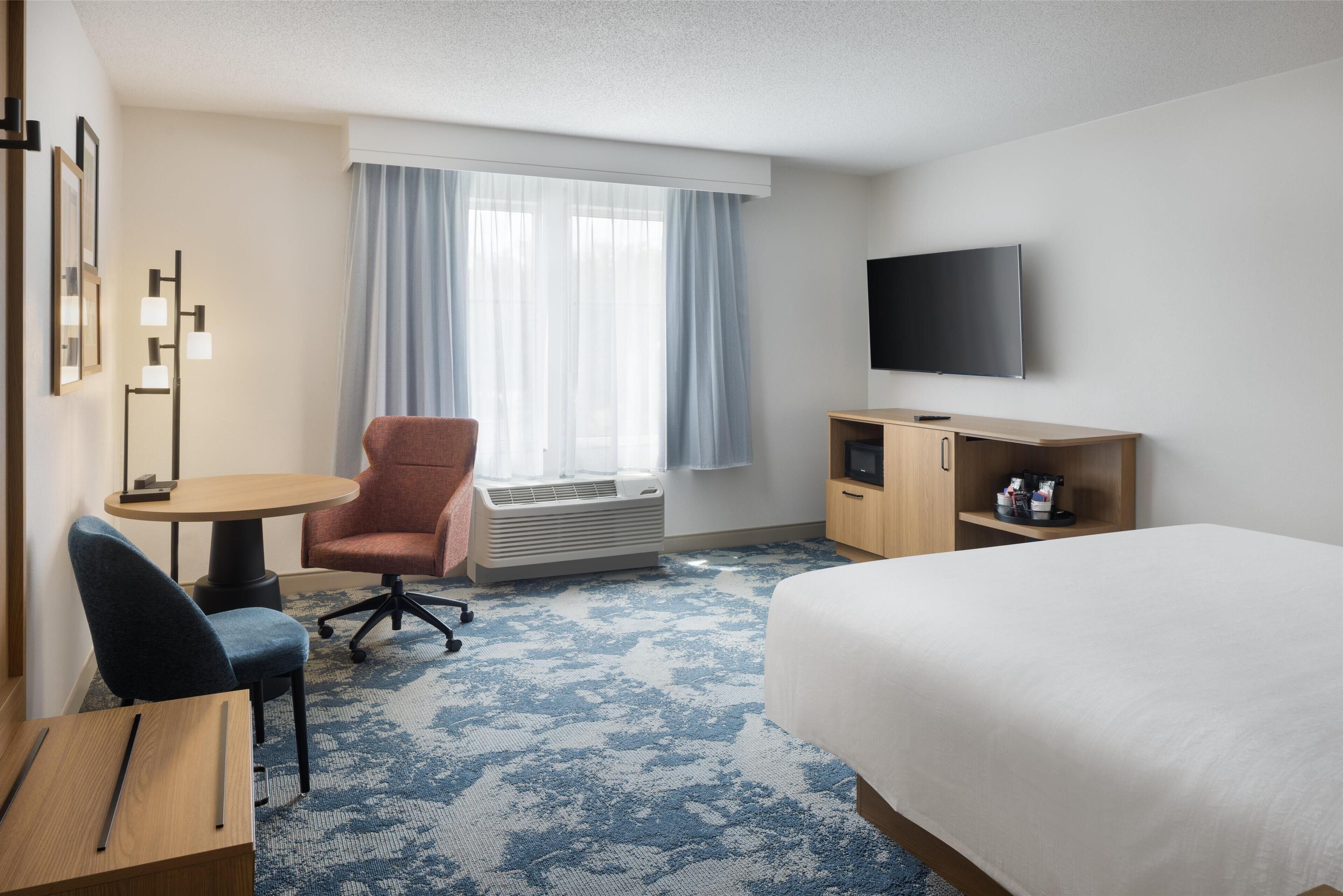 A hotel room with a bed, a desk with a chair, a television, a small cabinet, and a window with curtains.
