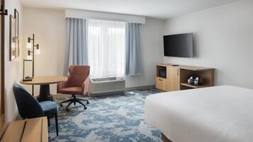 A hotel room with a bed, a desk with a chair, a television, a small cabinet, and a window with curtains.