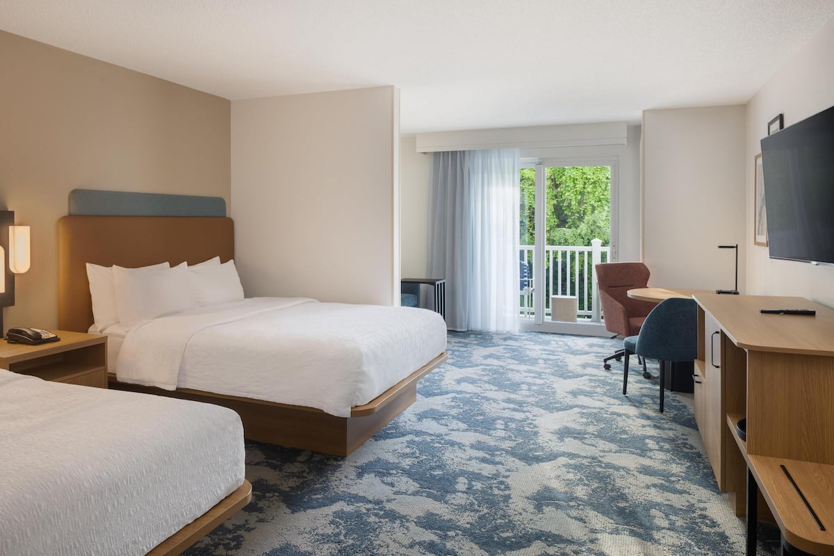 A hotel room with two beds, a desk, a chair, a TV, and a balcony with a view of greenery.