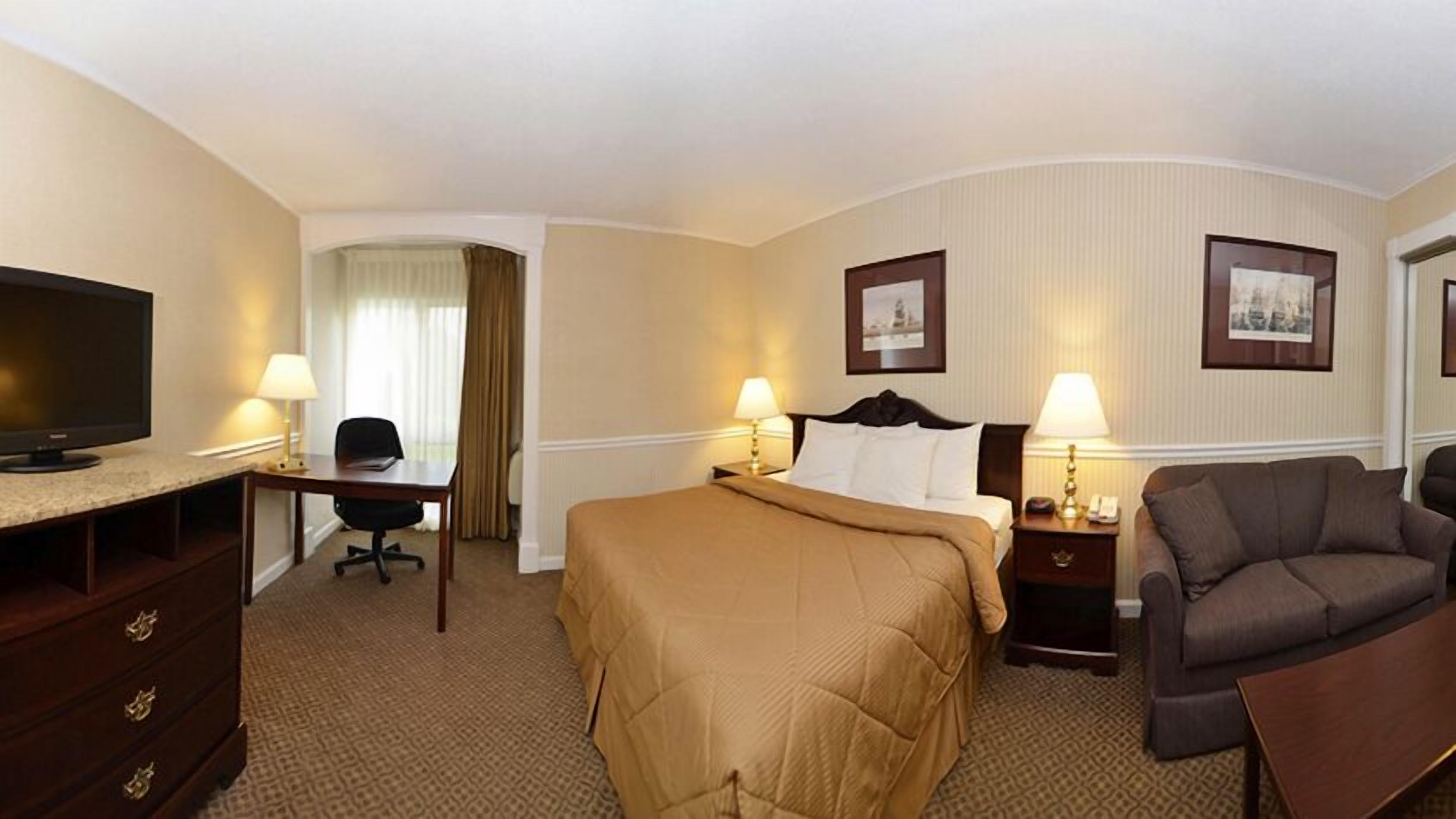 A hotel room with a bed, a desk with a chair, a television, two bedside lamps, a nightstand with a lamp, a sofa, and two framed pictures on the wall.