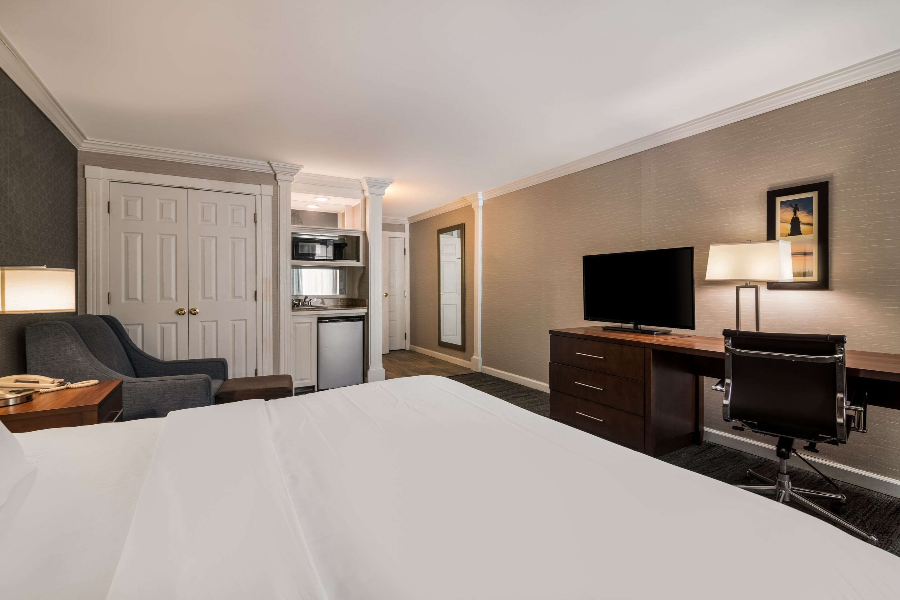 A hotel room with a bed, desk, chair, TV, lamp, and kitchenette.