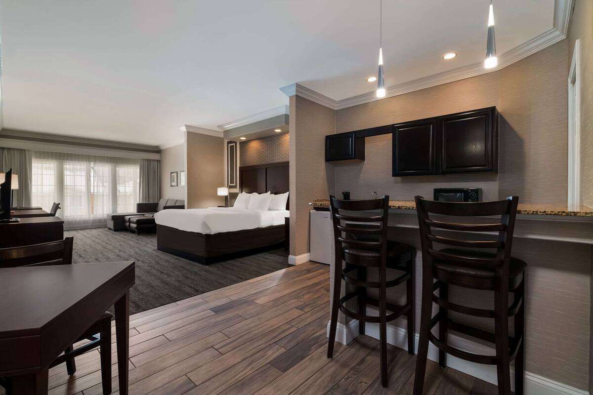 A hotel room with a bed, a desk, two bar stools, a kitchenette, and a TV.