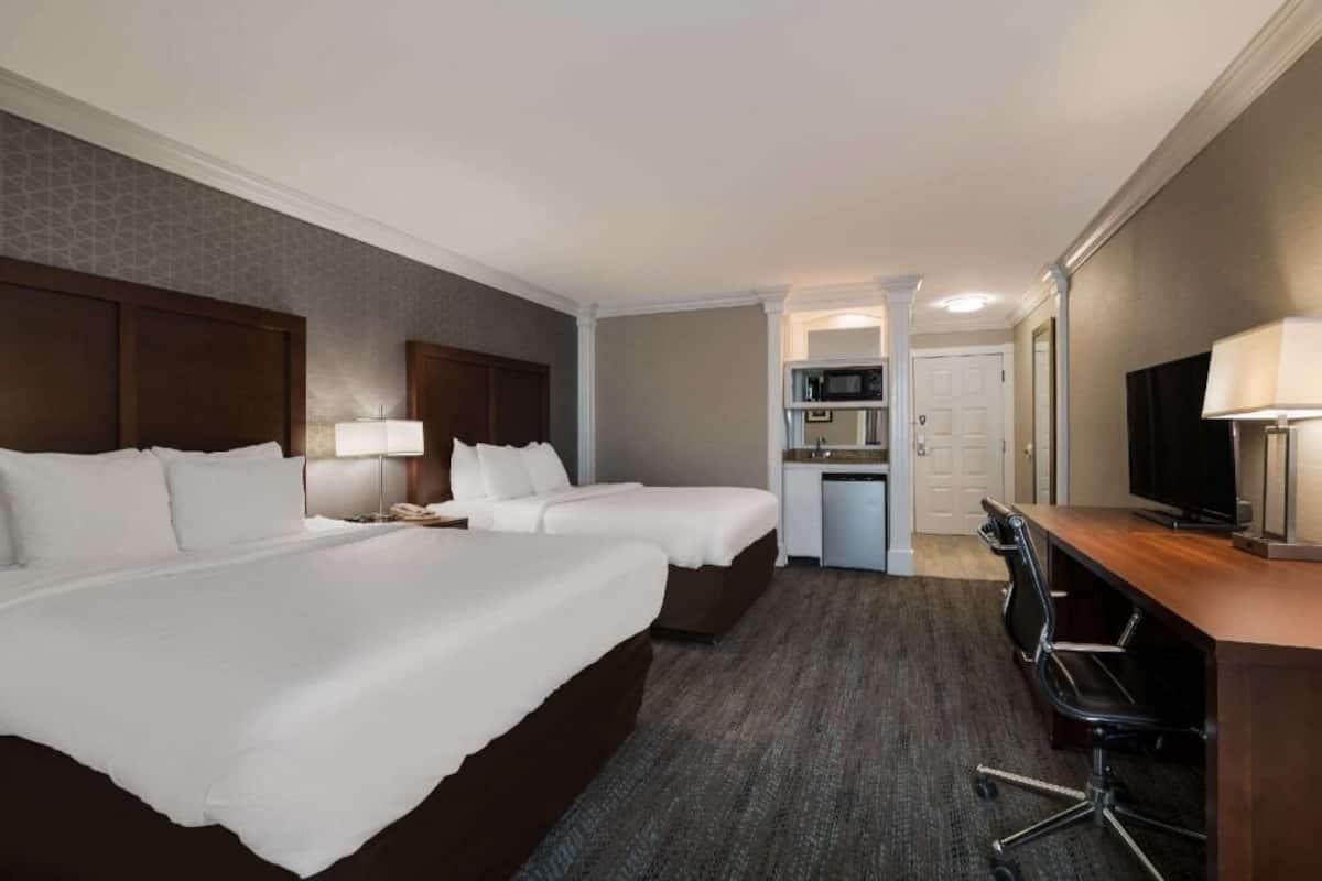 A hotel room with two beds, a desk, a TV, and a kitchenette.
