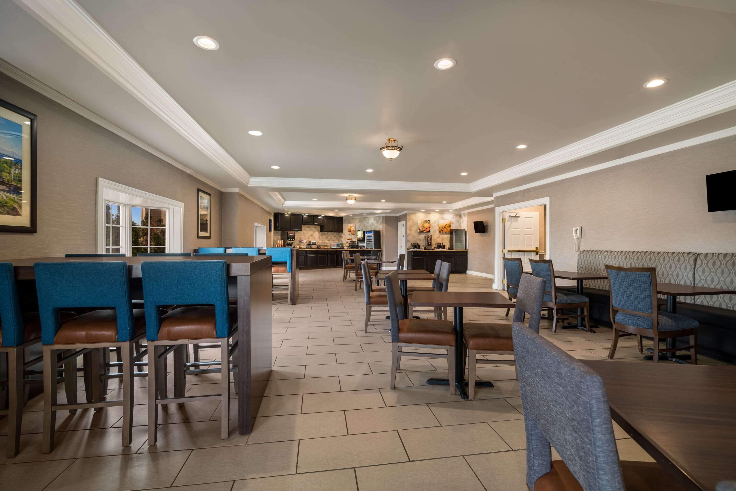 A spacious dining area with multiple tables and chairs, a kitchen in the background, and a ceiling with recessed lighting.