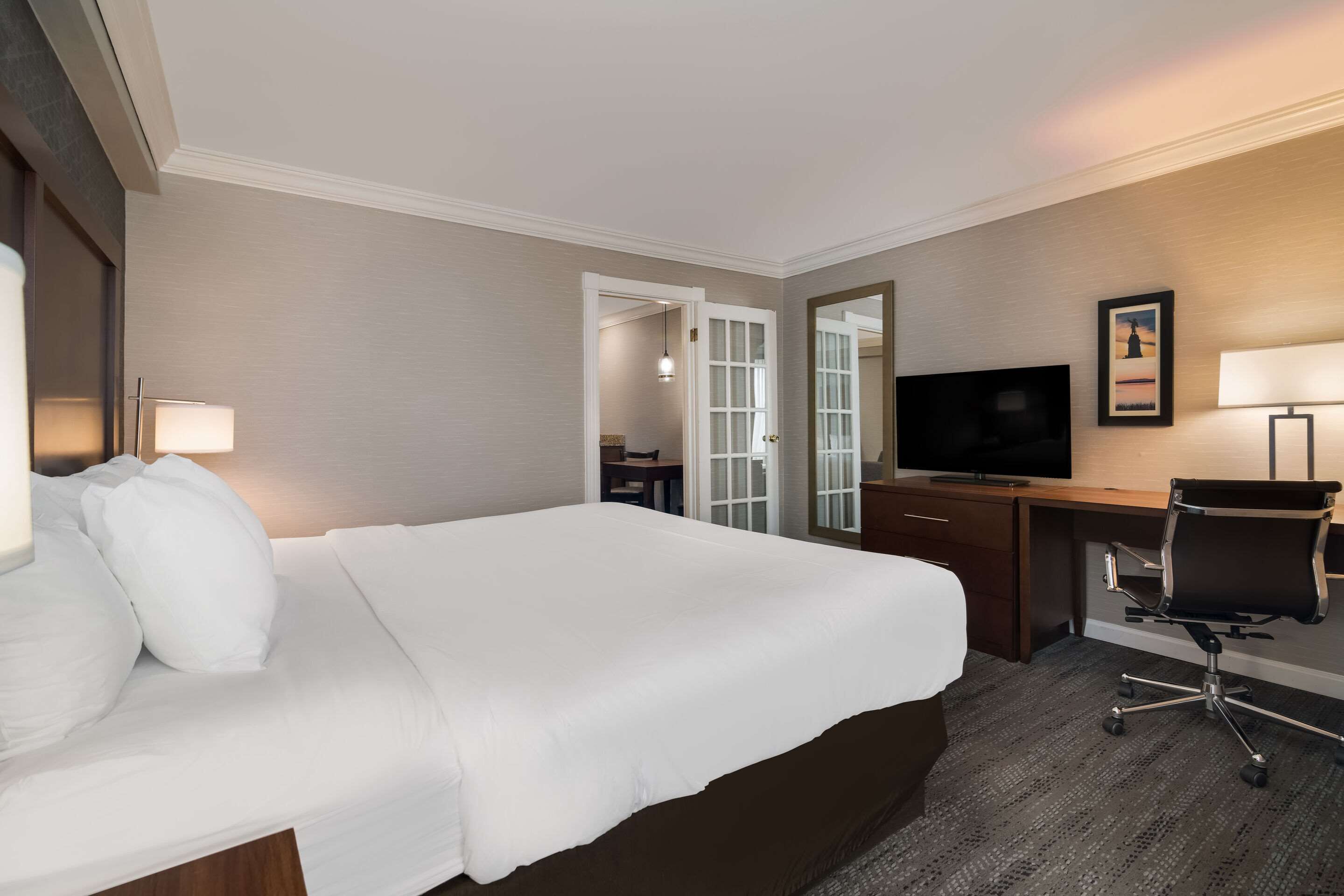 A hotel room with a large bed, a desk with a chair, a television, and a mirror.