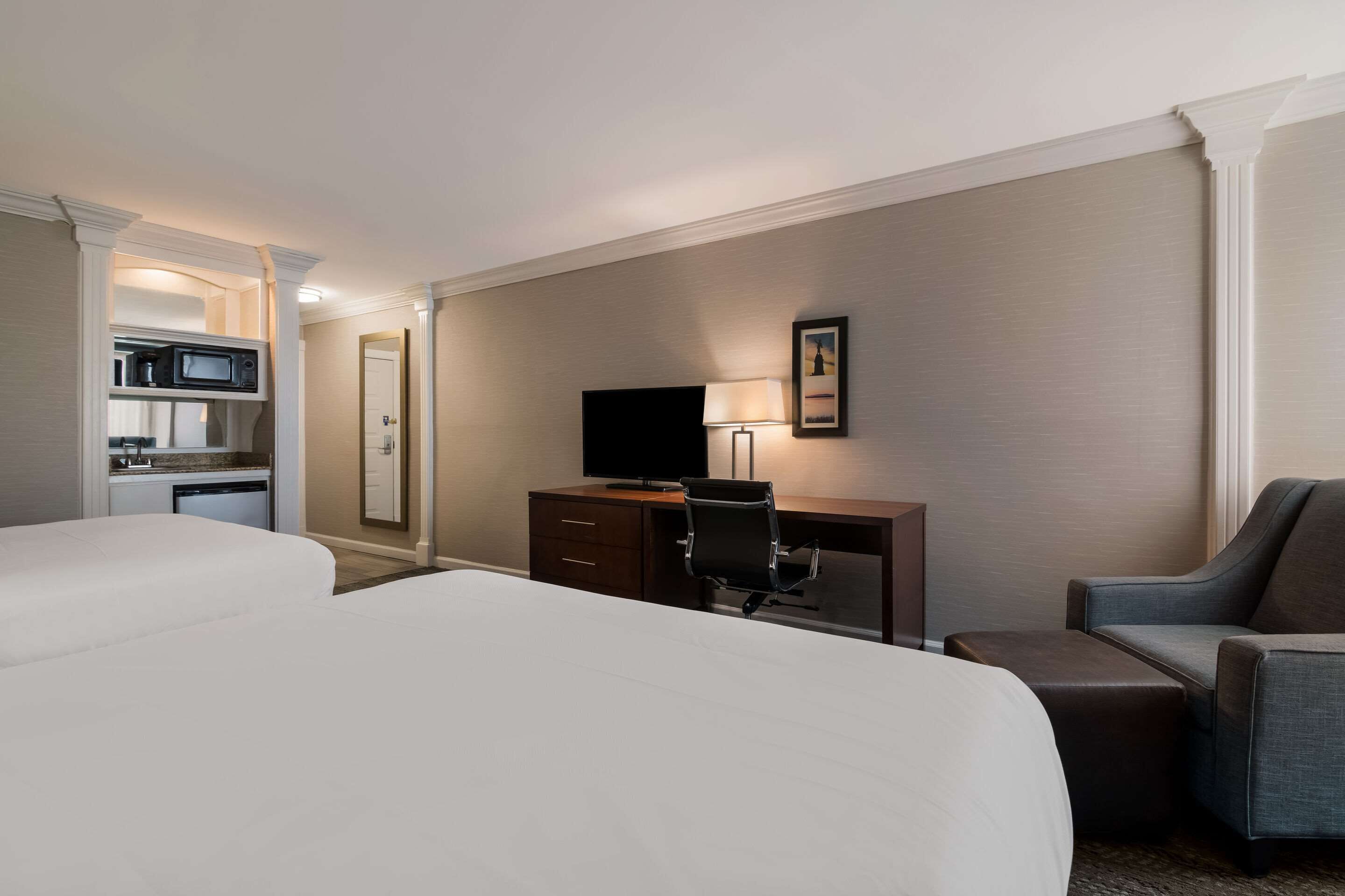 A hotel room with two beds, a desk with a chair, a television, and a small kitchenette.