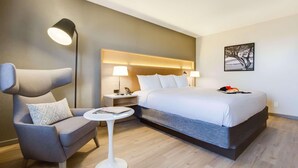 Una habitación de hotel moderna con una cama grande, un sillón gris, una mesita redonda y una lámpara de pie.