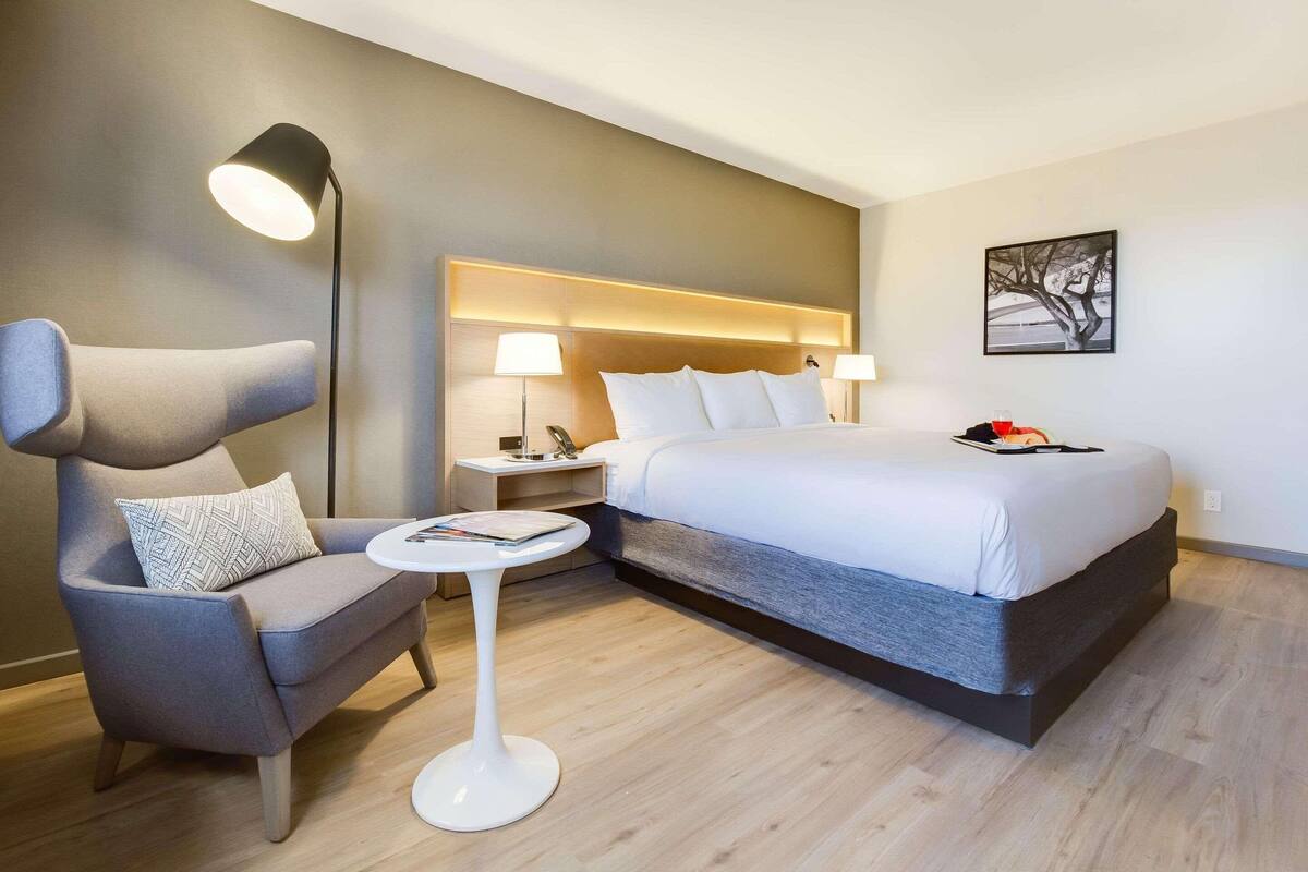 Una habitación de hotel moderna con una cama grande, un sillón gris, una mesita redonda y una lámpara de pie.